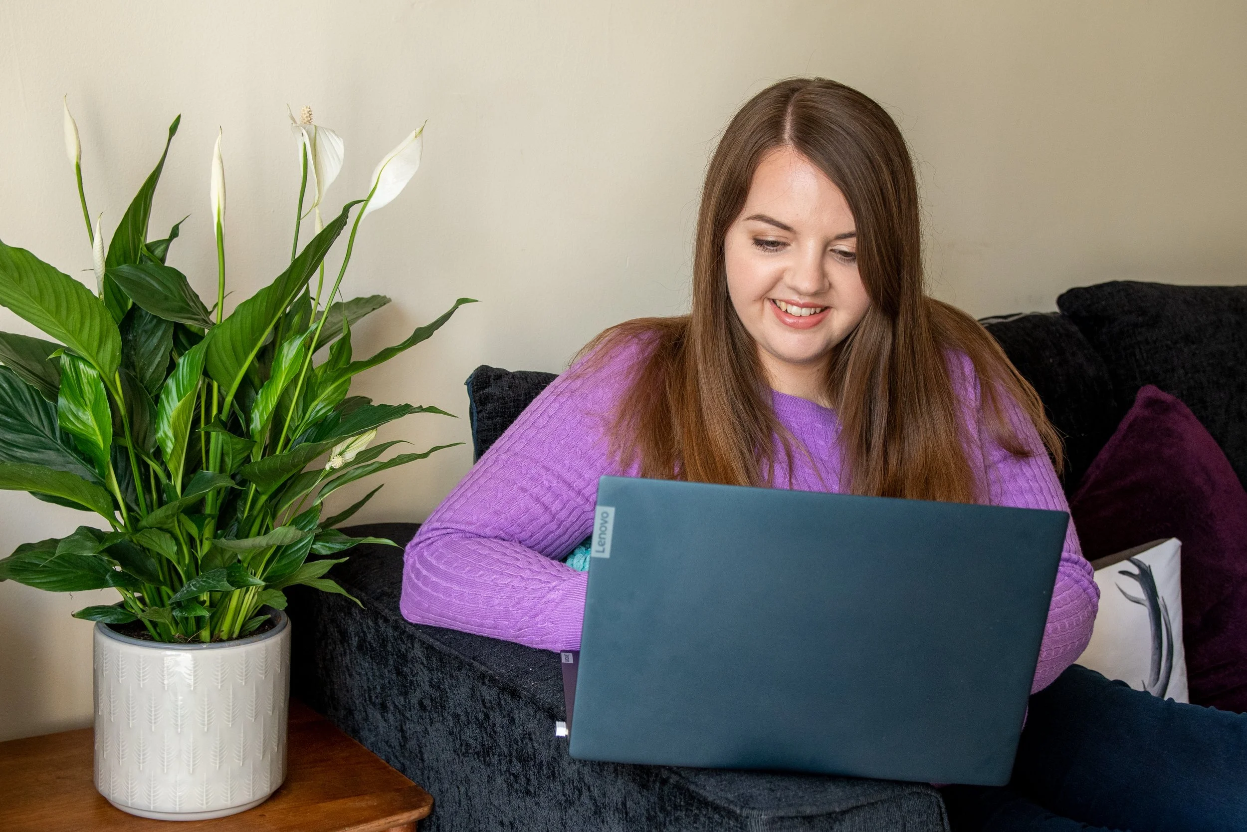 A young woman with long brown hair smiling and looking at a blue laptop. She is wearing a purple sweater and is seated on a black couch. To her left, there is a large potted plant with green leaves and white flowers. The background is a plain light-colored wall.