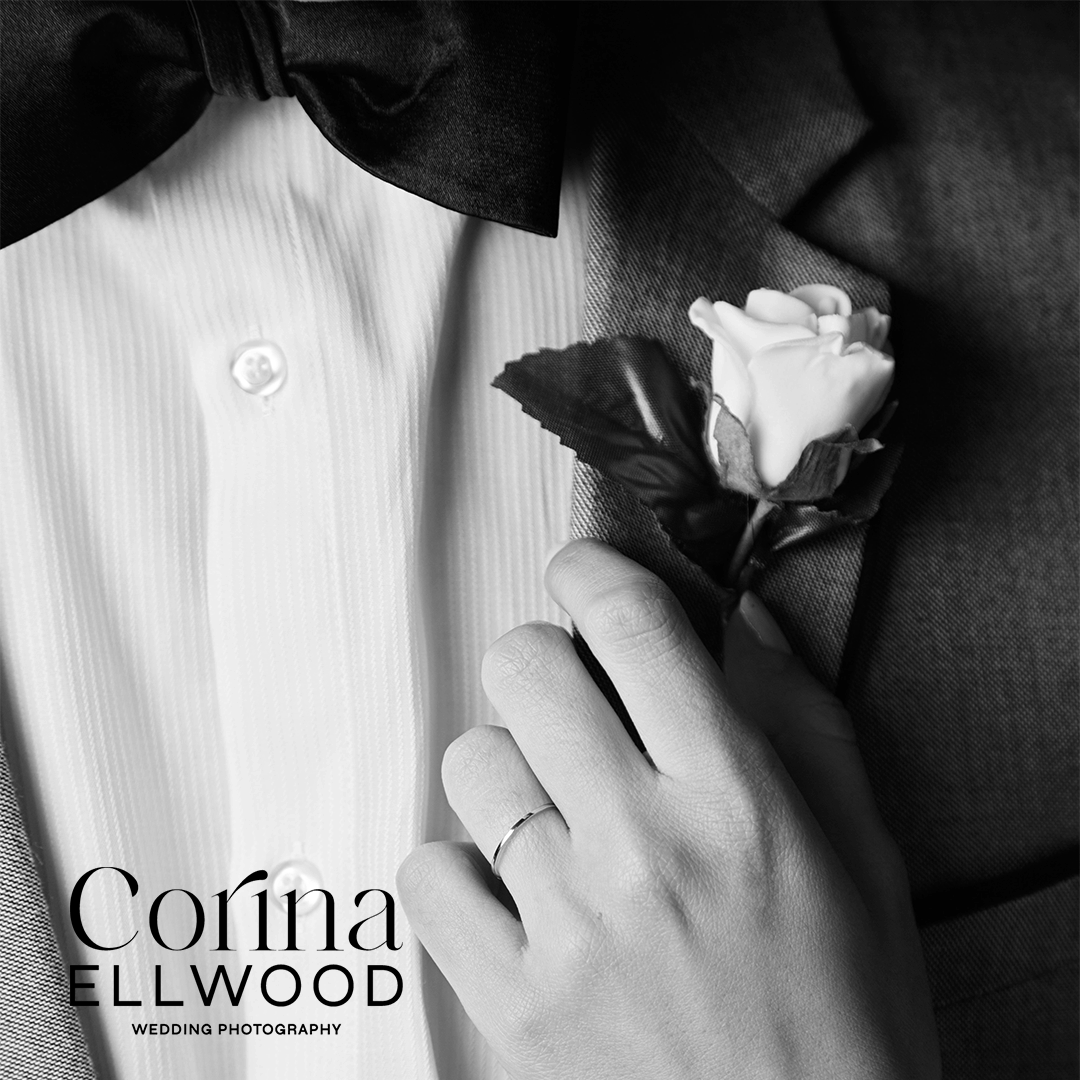 Close-up of a person's hand with a wedding ring, holding a white flower boutonniere on a tuxedo, with a white shirt and black bow tie visible.