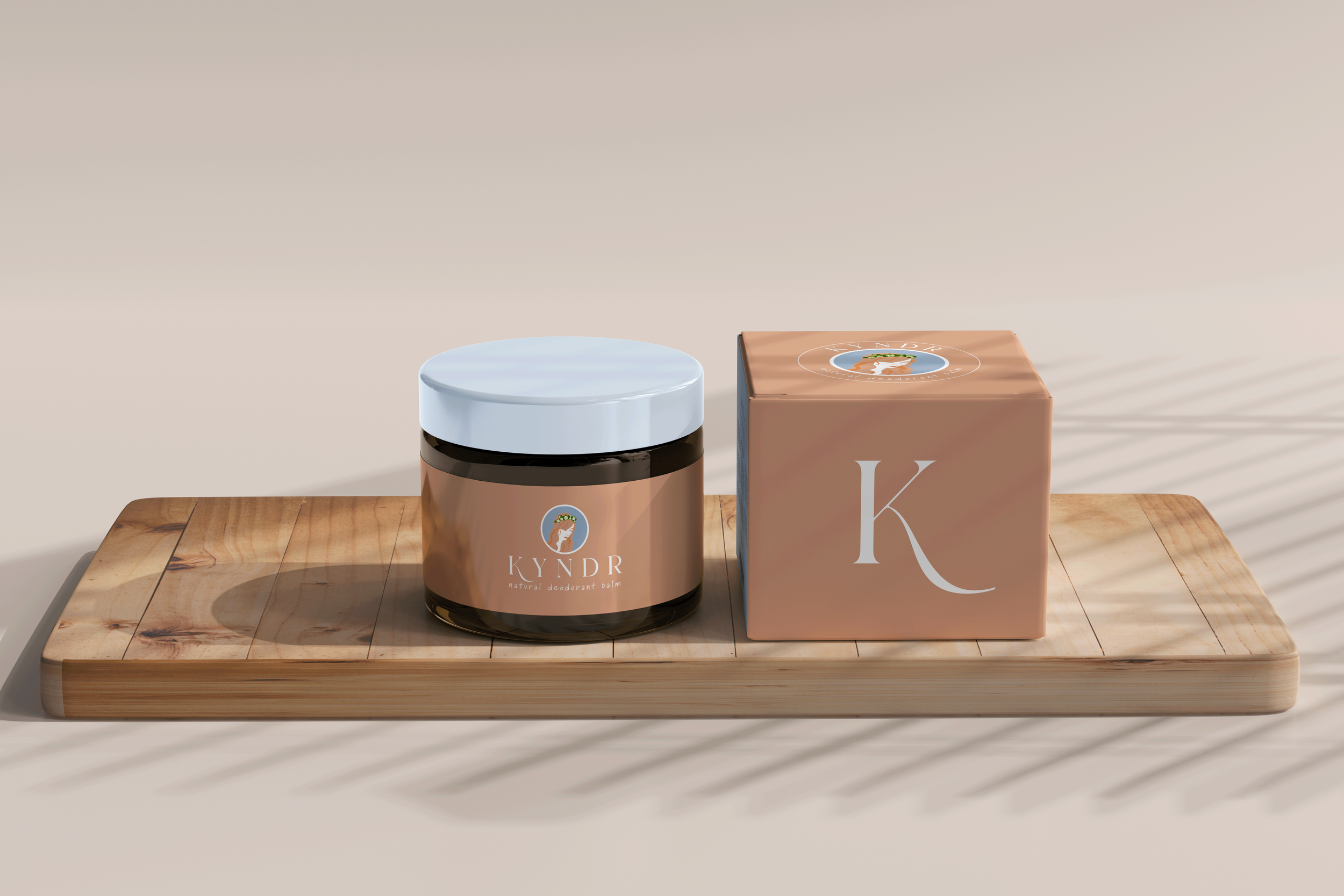 A jar of KYND natural deodorant balm and a pink box with a large white 'K' on it, placed on a wooden tray.
