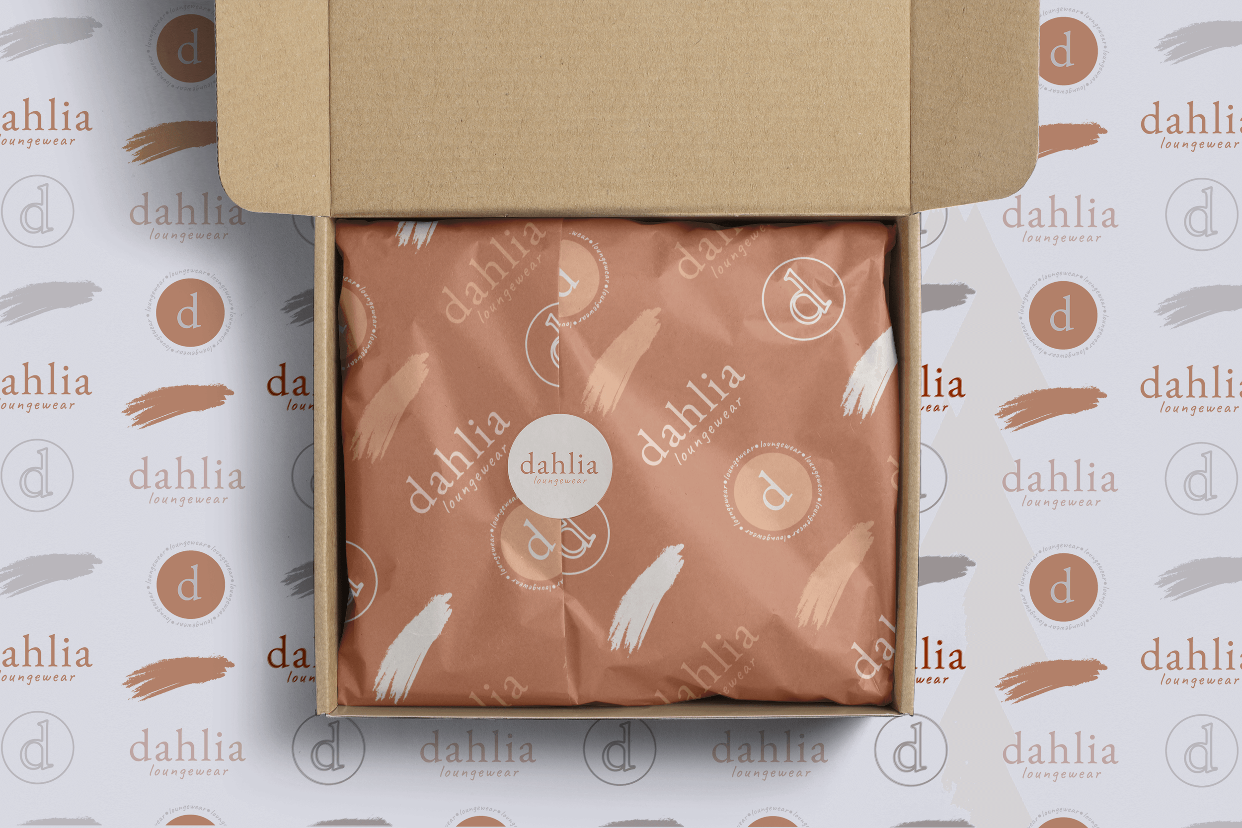 Open cardboard box containing wrapped product with Dahlia Loungewear branding, placed on patterned wrapping paper with Dahlia Loungewear logos and brushstroke designs.