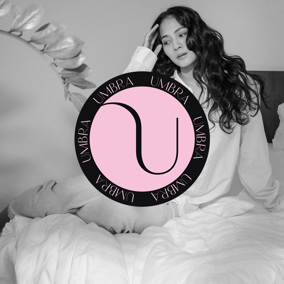 A woman with long, curly hair sitting on a bed with a thoughtful expression, touching her temple, black and white photo with a circular pink and black logo with the word 'UMBRA' repeated around it overlaid on the image.