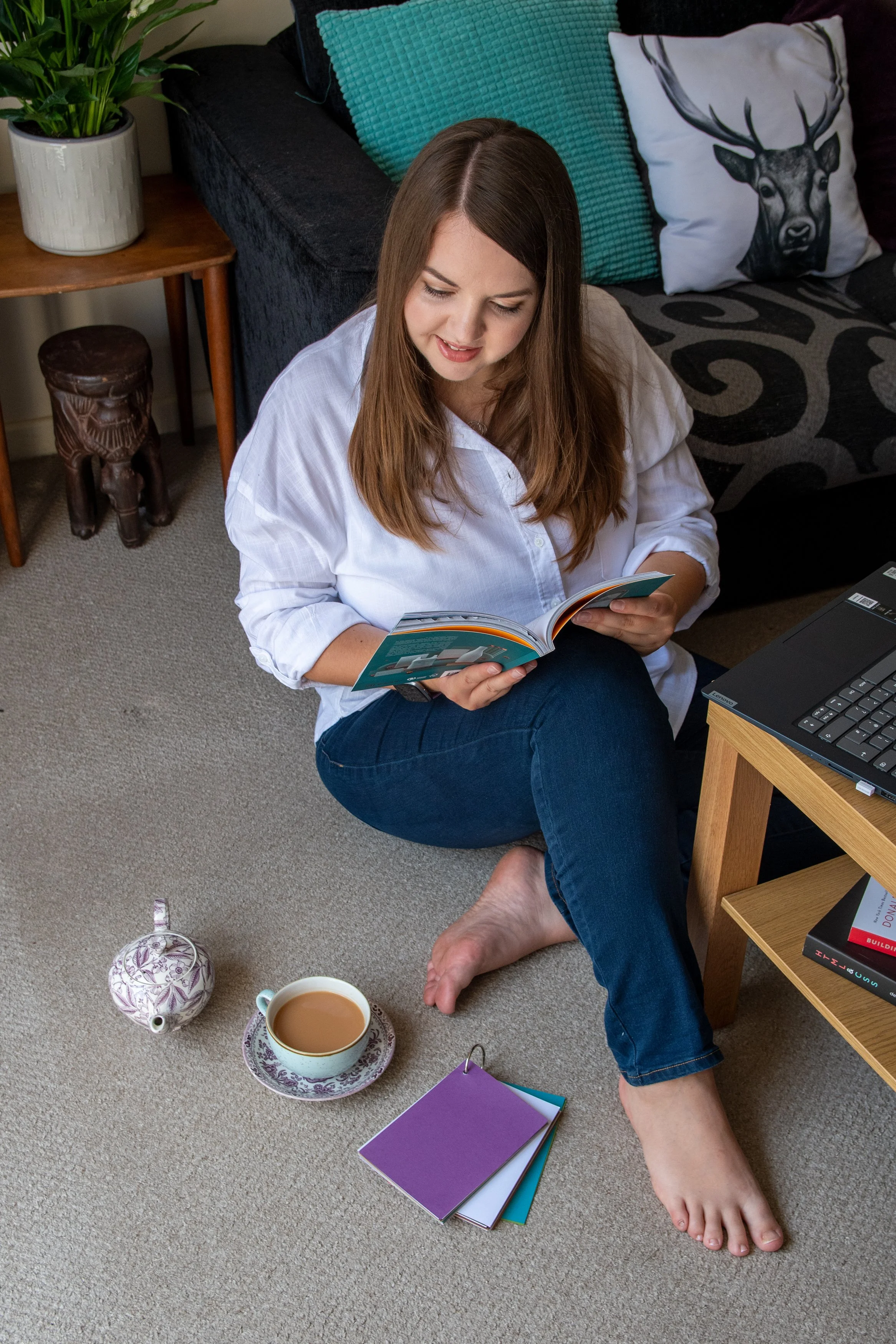 A woman with long brown hair is sitting on the beige carpet floor, reading a book. She is wearing a white shirt and blue jeans, and she is barefoot. Next to her on the floor are a purple and a green notebook. In front of her, there is a cup of coffee or tea on a saucer, and a white teapot with a floral design. To her left, there's a wooden side table with a potted green plant. Behind her, there's a black couch with turquoise and animal print pillows, one of which has a deer head illustration.