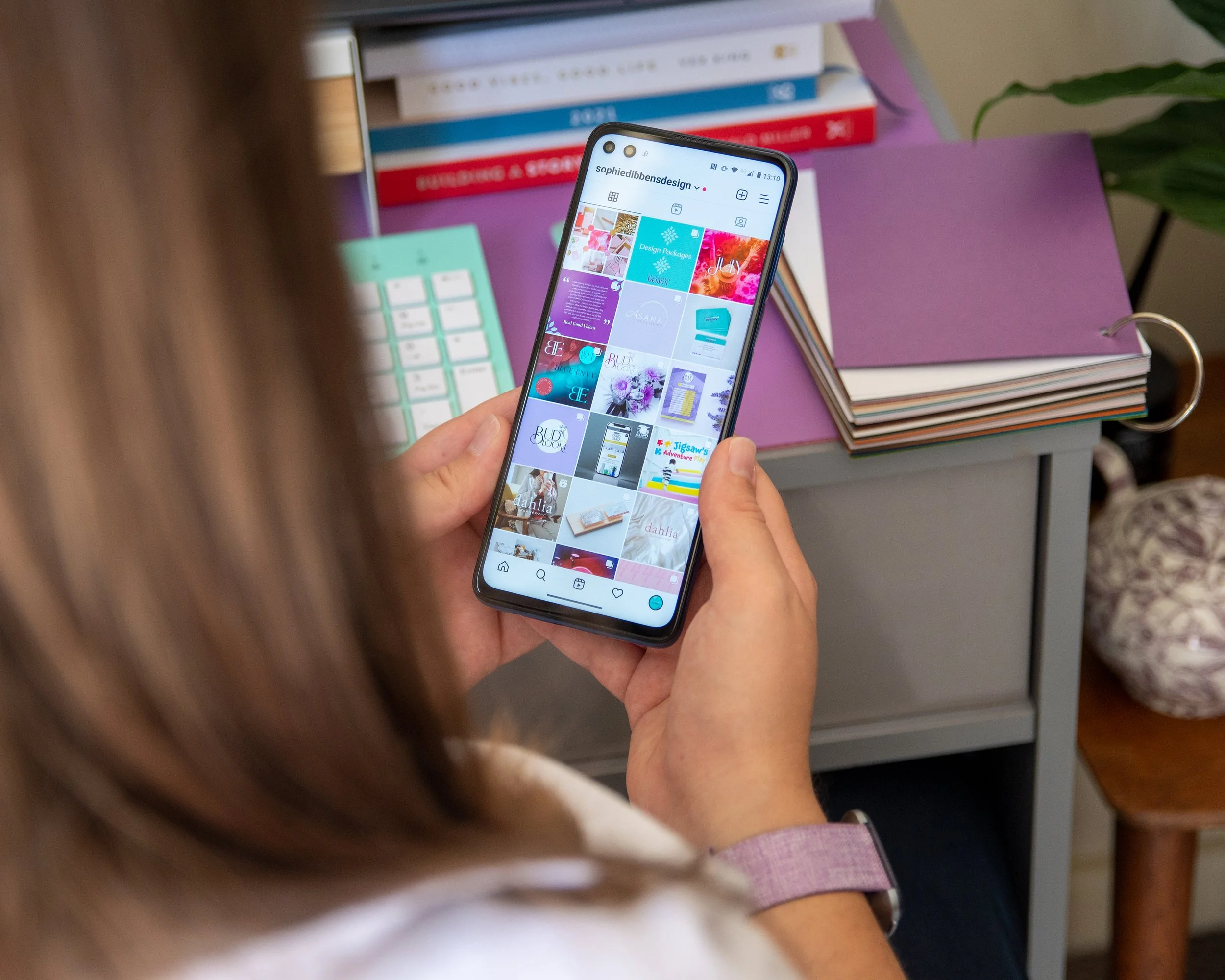 Person holding a smartphone displaying a social media app or photo gallery, with books and a purple notebook in the background.