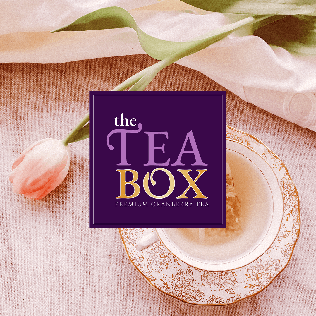 A cup of cranberry tea on a floral saucer, with a pink tulip and a purple and gold tea box labeled 'The Tea Box Premium Cranberry Tea' nearby, on a pink textured surface.
