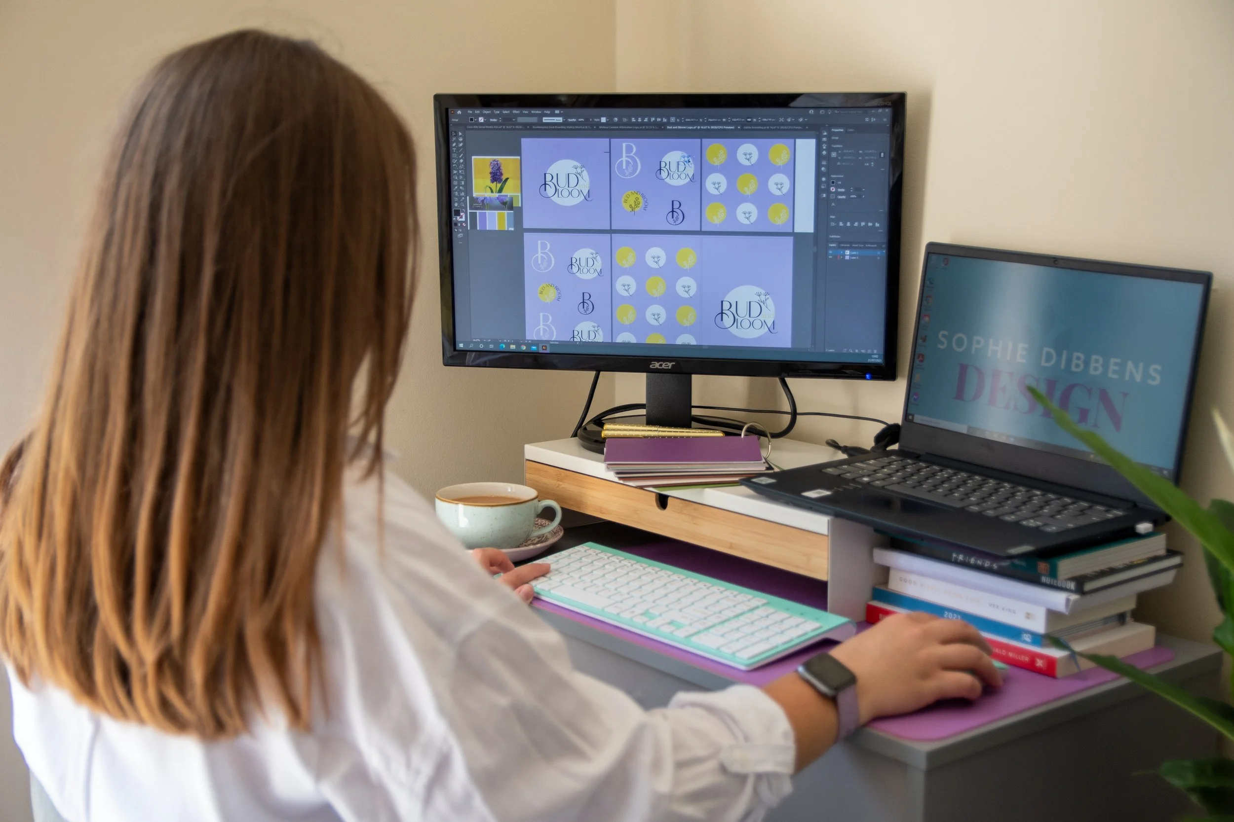 A woman with reddish-brown hair working at a desk, using a computer with design software open. The desk has a pink mat, a white keyboard, a mouse, a cup, and stacks of books. Two monitors are visible, one displaying a logo and design elements, and the other showing the text 'Sophie Dibbens Design.'