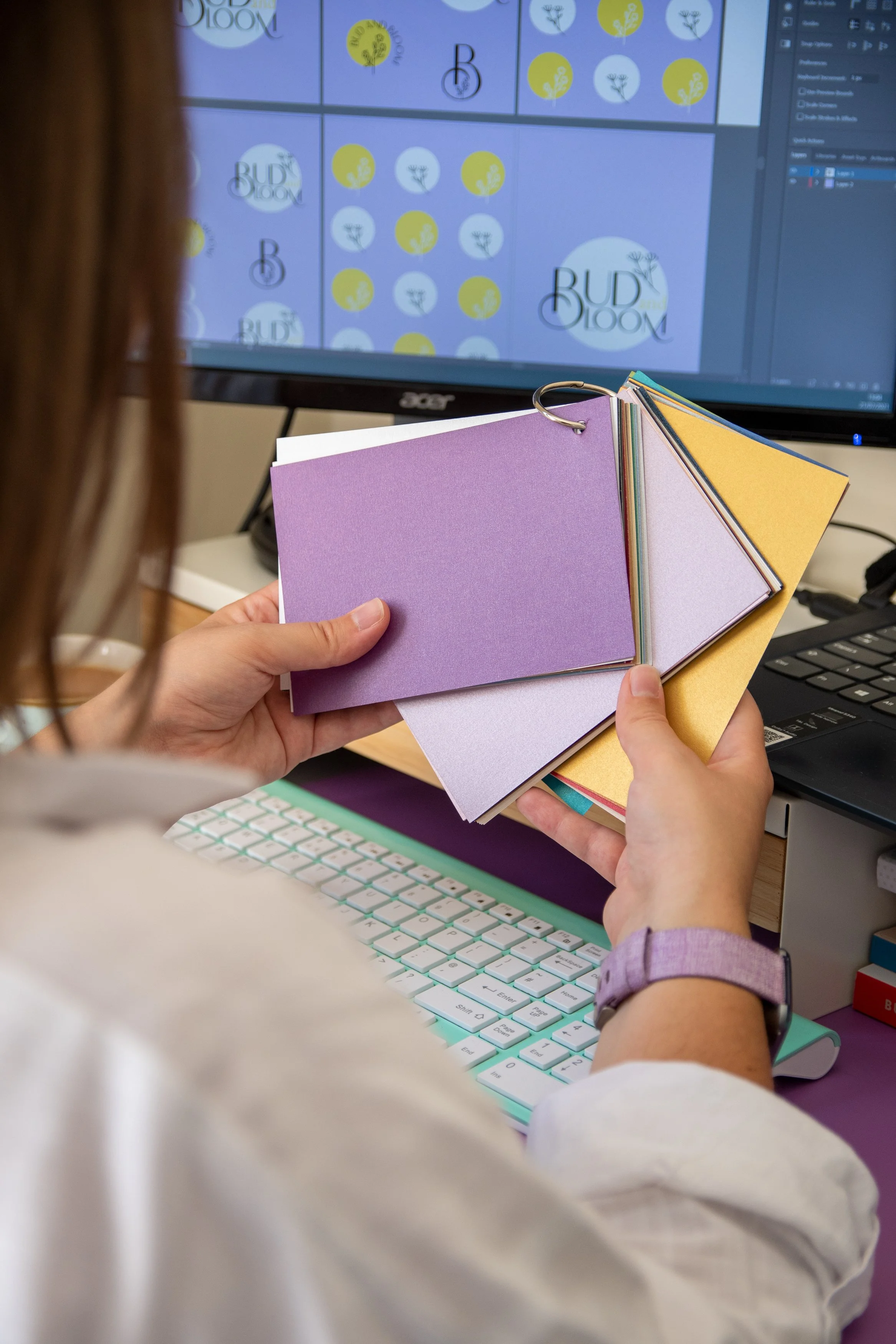 A person holding a color fan with purple, white, yellow, and pink paper samples in front of a computer monitor displaying a design with the words 'BUD BLOOM' and multiple yellow and white flower logos. The person is sitting at a desk with a keyboard and a computer mouse.