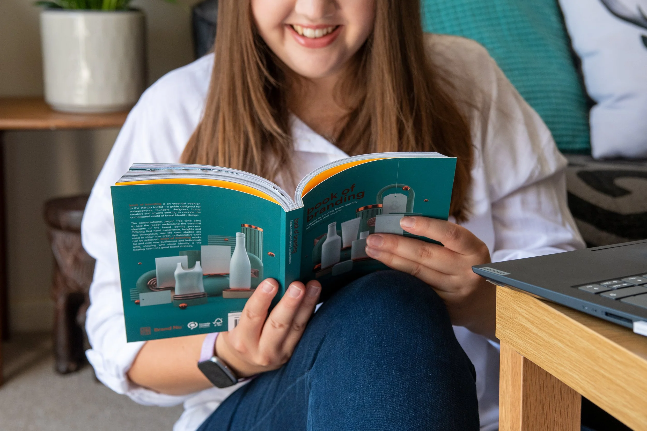 A woman with long brown hair is smiling while reading a book titled 'Book of Branding'. She is sitting at a desk with a laptop nearby, wearing a white shirt and blue jeans.