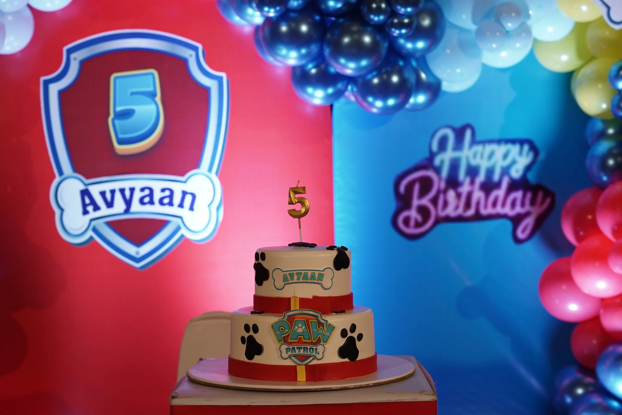 A birthday scene with a cake decorated with Paw Patrol logos and black paw prints, placed on a table. In the background, colorful balloons in blue, white, yellow, and pink fill the scene. A sign with the number 5 indicates a fifth birthday celebration. There is also a sticker or topper with the name 'Ayyan' on the cake.