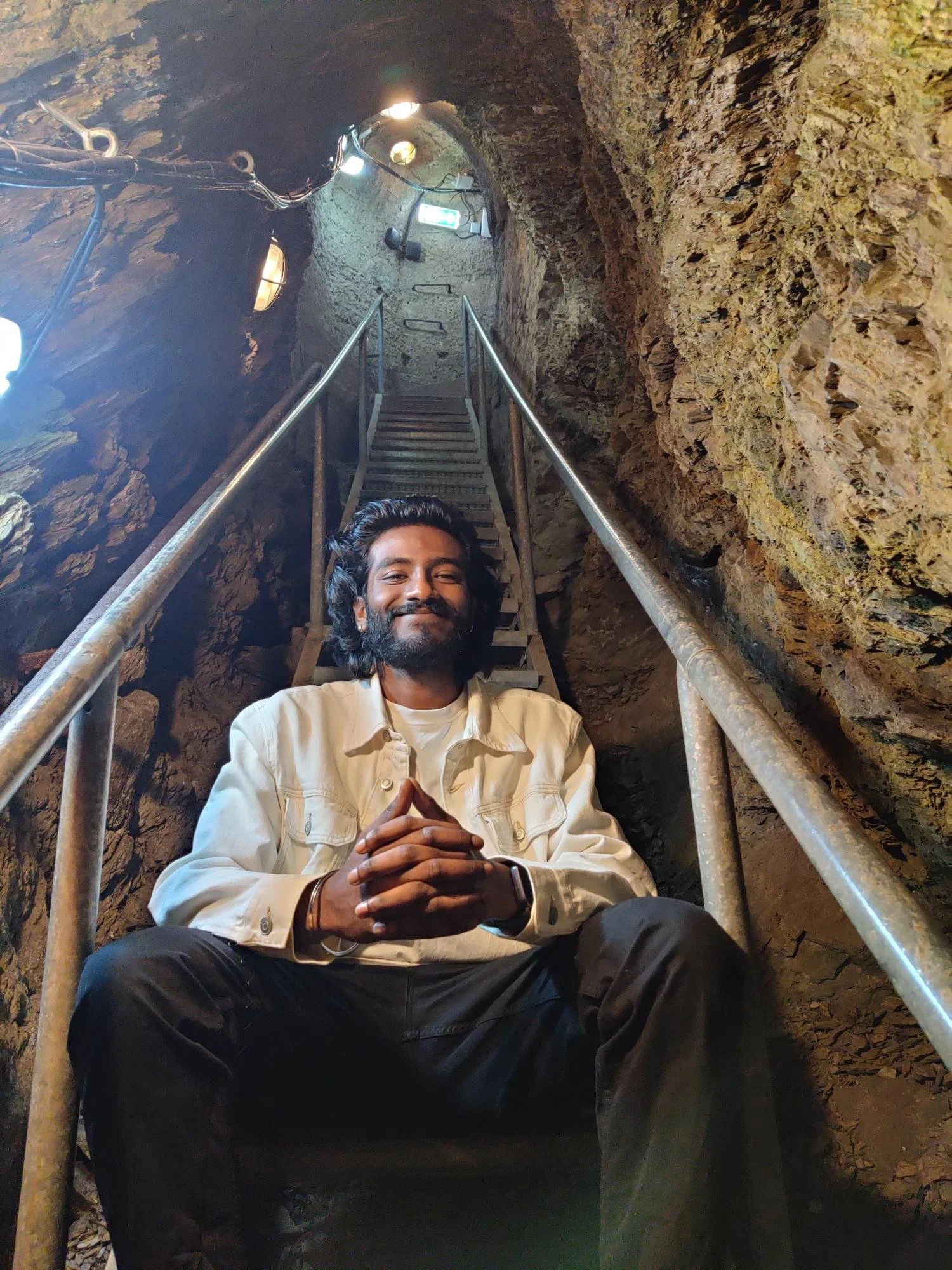 A man sits on a staircase inside a narrow tunnel with rocky walls, smiling and clasping his hands together. The tunnel has metal handrails and opening lights.