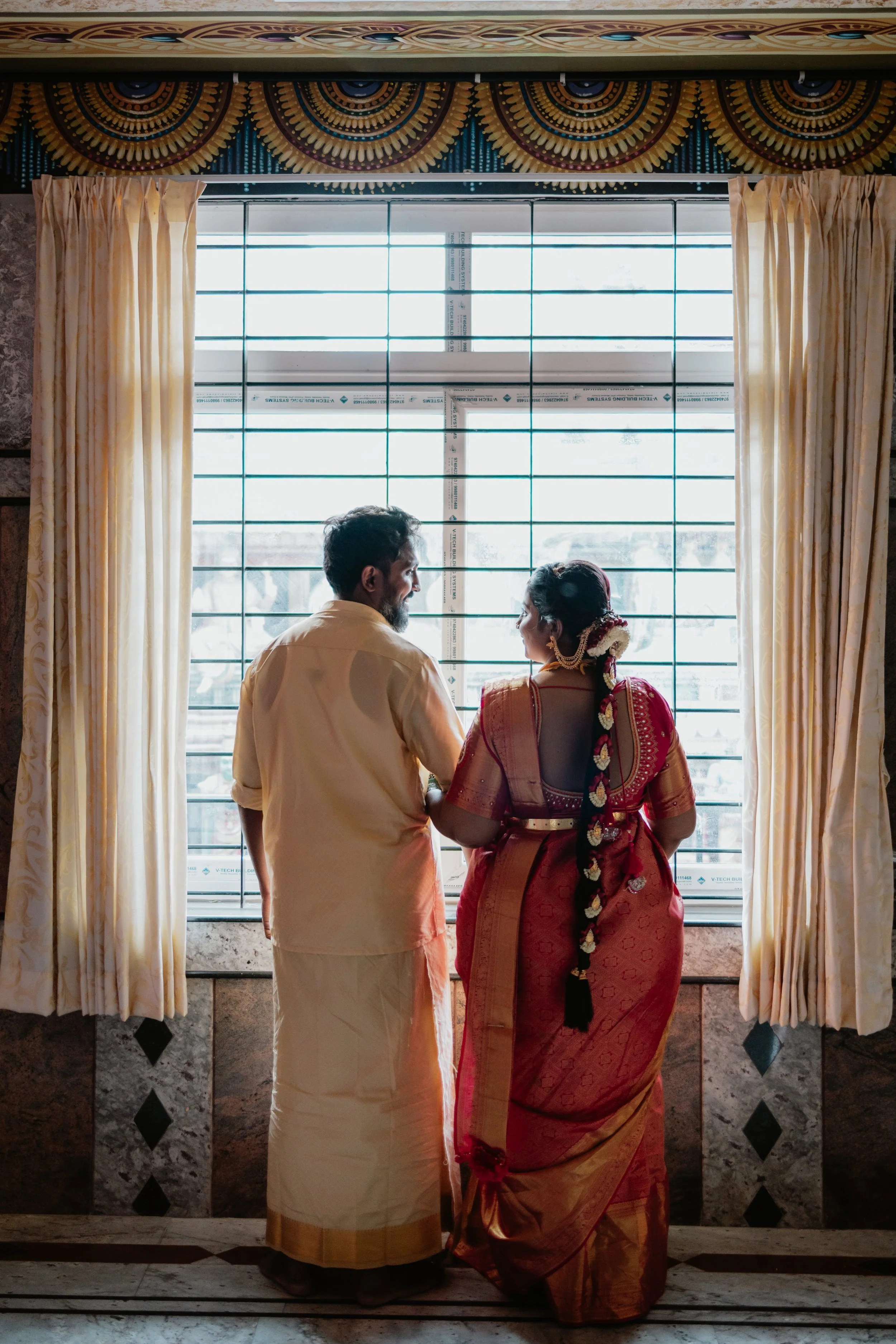 A man and woman dressed in traditional Indian attire standing close together near a window with cream-colored curtains, viewed from behind.