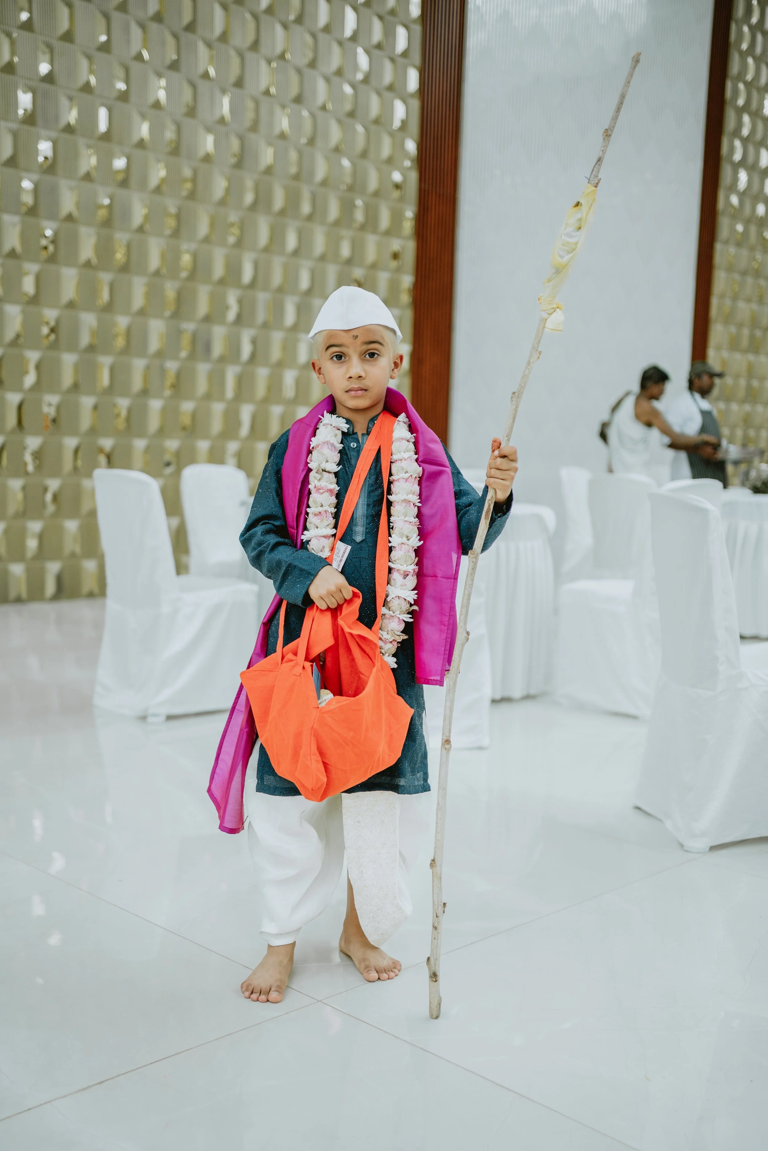 A young boy dressed in traditional Indian attire, holding a stick with a cloth tied on top. He is wearing a white cap and a flower garland around his neck, with a pink cloth draped over his shoulder. The background shows a decorated indoor setting with round tables covered with white tablecloths and two people in the background.