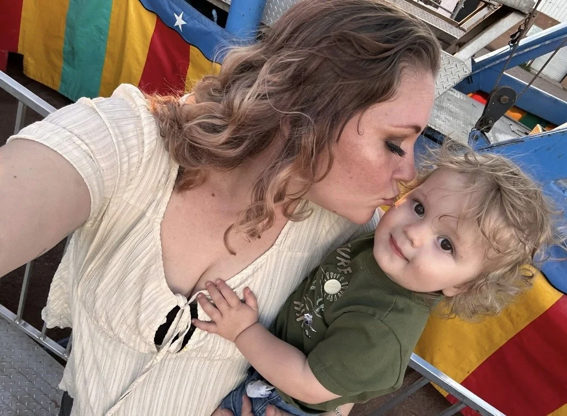 A woman and a young child sharing a tender moment at an amusement park or fair, with the woman kissing the child's cheek. The woman has wavy brown hair, and the child has curly blonde hair, wearing a green shirt. In the background, there is a colorful carnival booth or ride with a blue, yellow, and red color scheme.