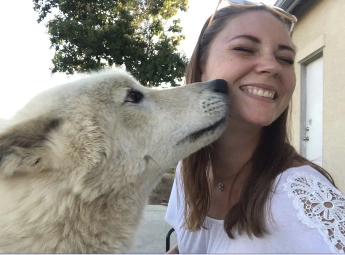 A young woman smiling as a light-colored dog, possibly a Labrador Retriever, licks her face outdoors.