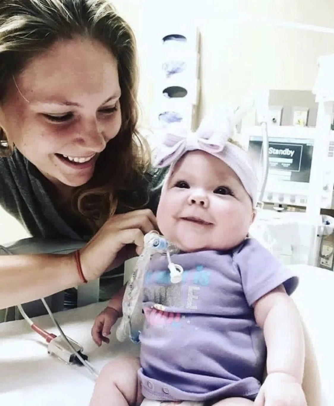 A smiling woman holding a baby girl with a pink bow headband and a pacifier clip, in a hospital setting.