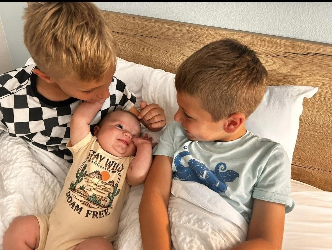 Three young boys lying in bed together, smiling at each other. The youngest is in the middle, and the two older boys are on either side, leaning over to look at the baby.