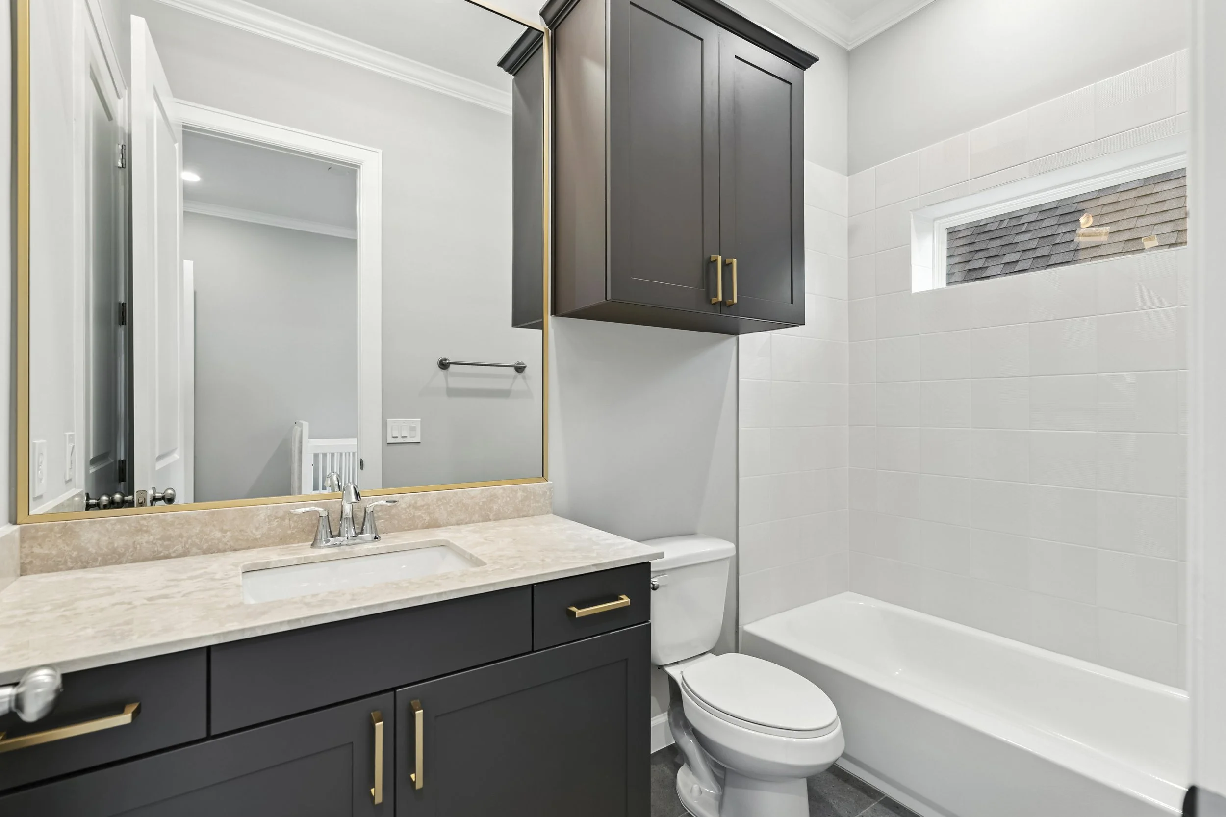Modern bathroom with navy vanity, beige marble countertop, large mirror with gold trim, white toilet, dark cabinet above the toilet, white tiled shower with small window.