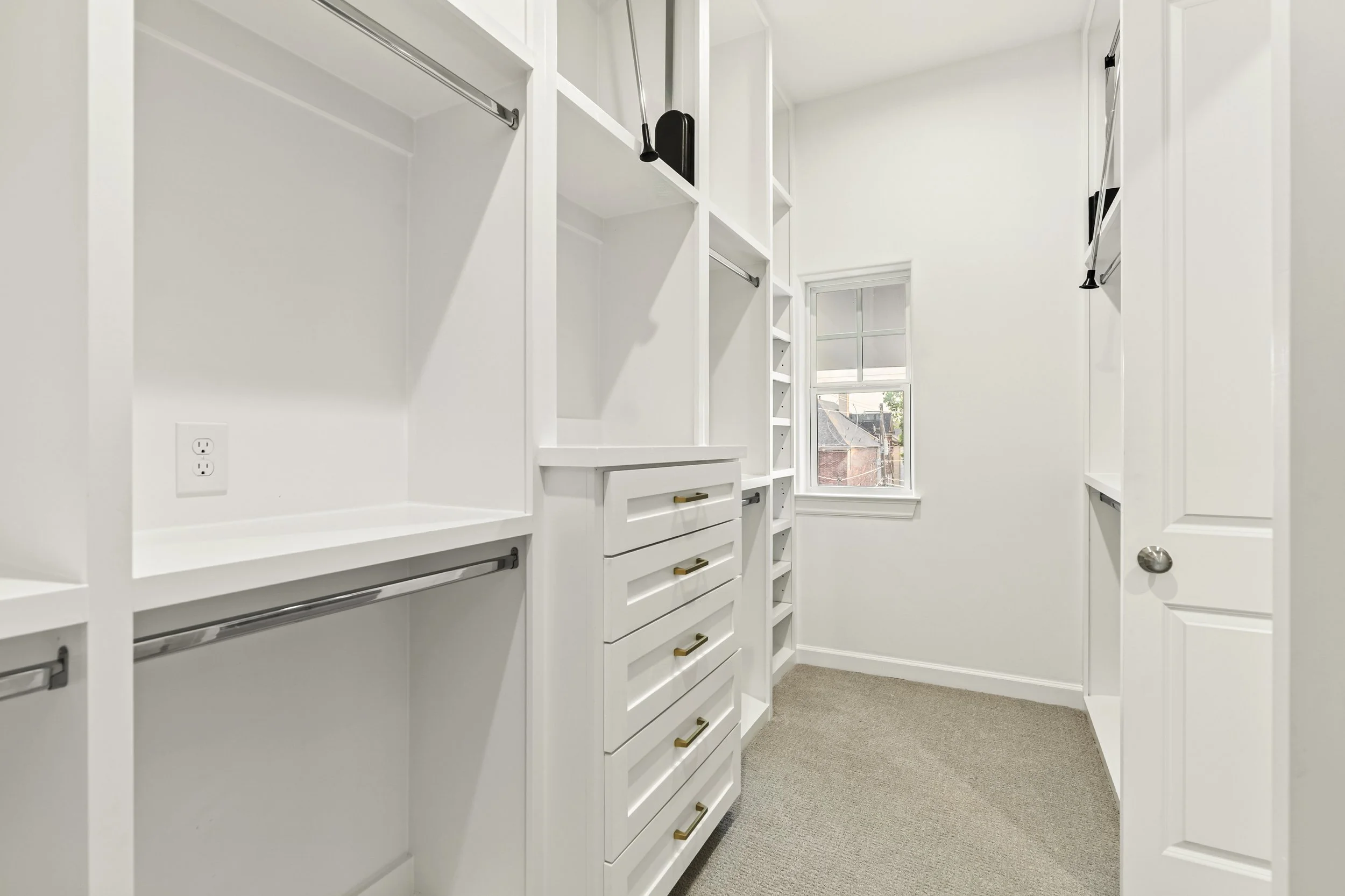 Empty white walk-in closet with built-in shelves and drawers, carpeted floor, window with view outside.