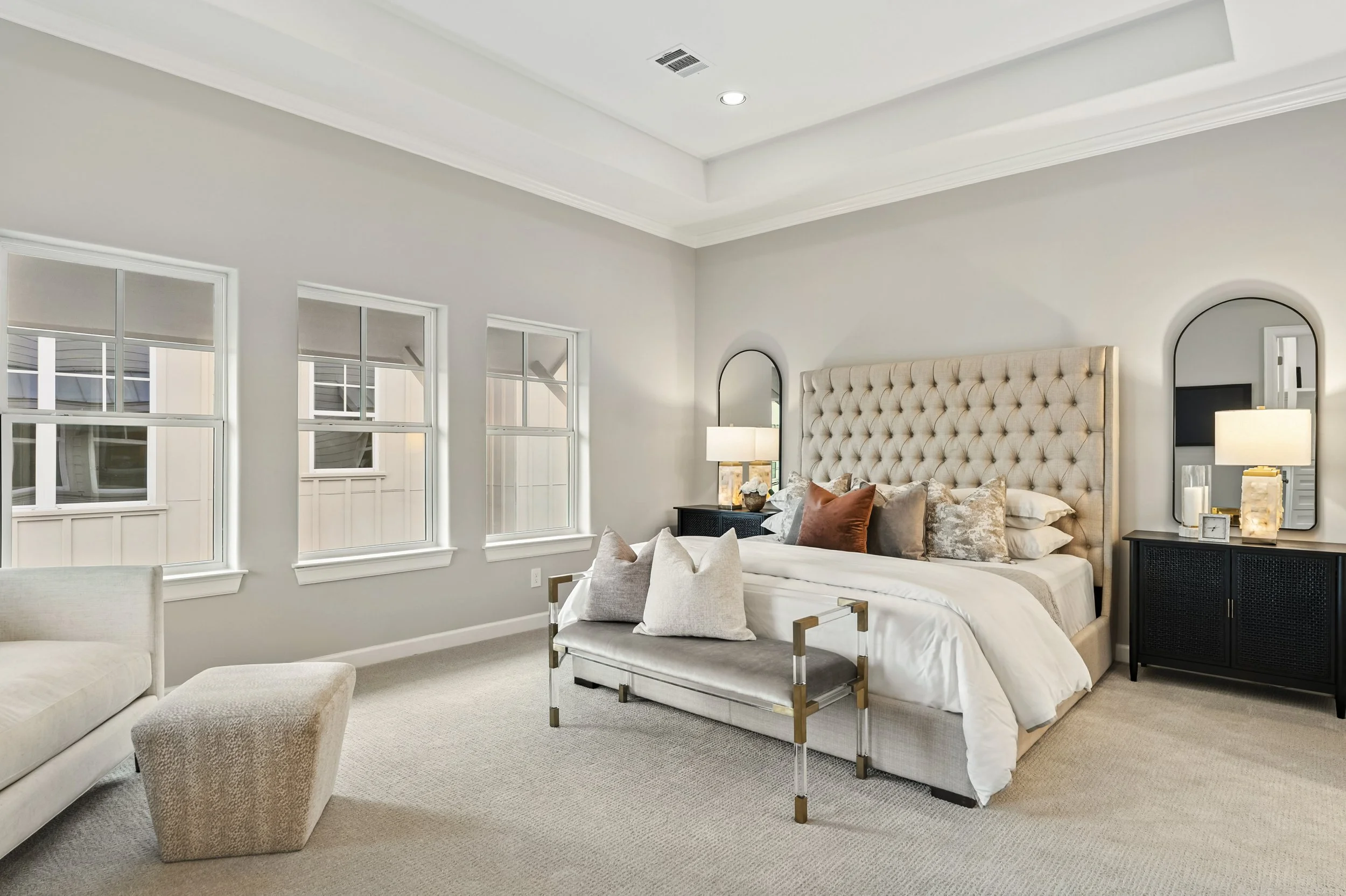 Modern bedroom with a large tufted headboard, nightstands with lamps, a bench at the foot of the bed, and multiple windows letting in natural light.