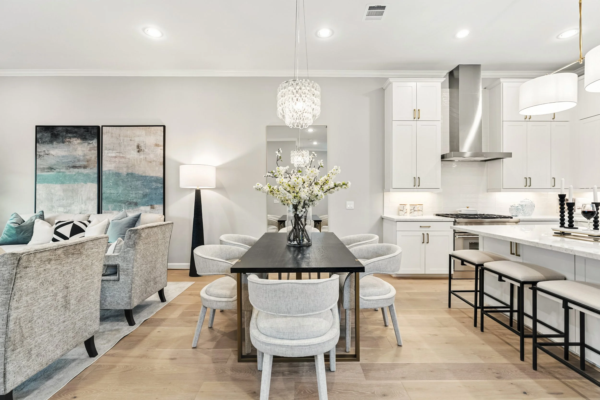 Open-concept living and dining area with a kitchen. The dining table has a vase with white flowers, surrounded by light-colored chairs. The living area has gray and cream sofas with pillows, art pieces on the wall, and a tall floor lamp. The kitchen has white cabinets, a stainless steel range hood, and black barstools at a white countertop. Pendant and chandelier lighting add to the bright, modern aesthetic.