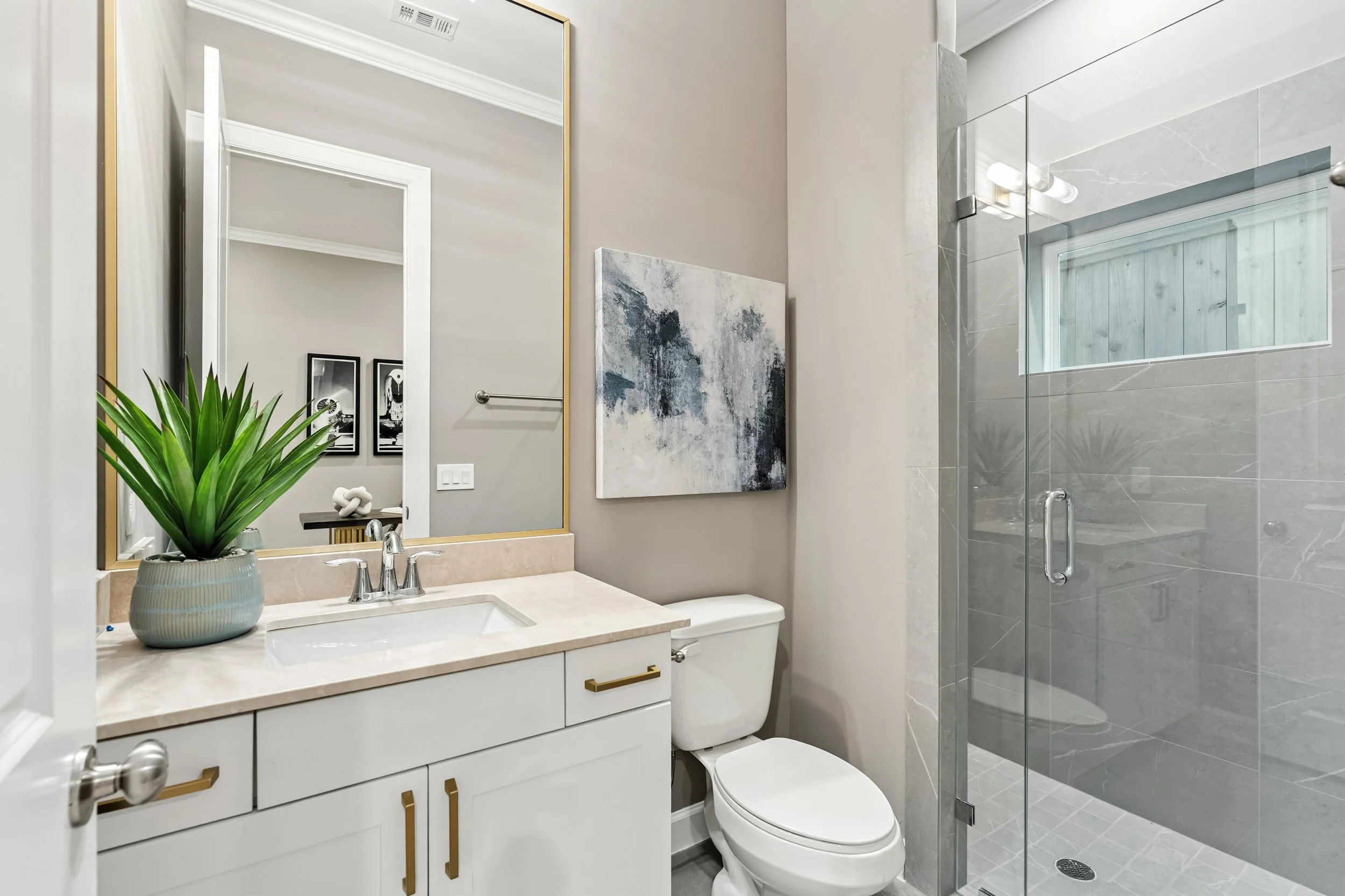 Modern bathroom with a large mirror, a plant on the vanity, abstract artwork, toilet, and glass-enclosed shower with gray tiles and a window.