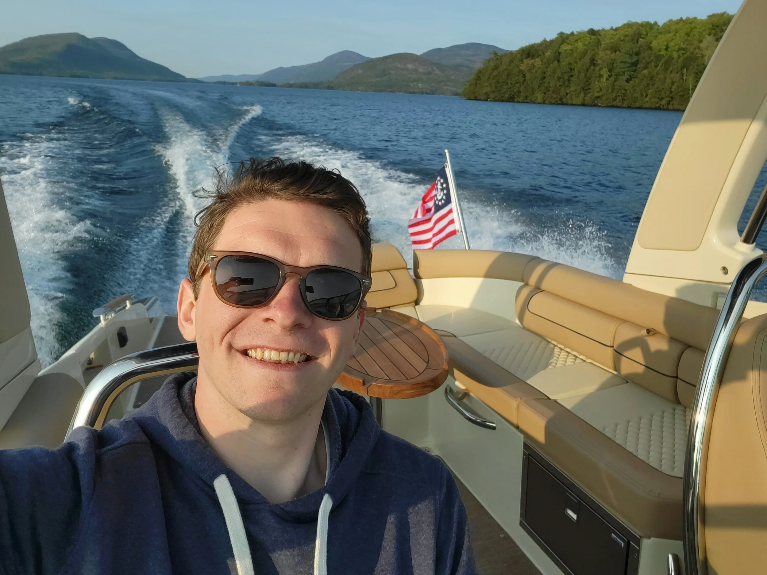 A smiling man wearing sunglasses taking a selfie on a boat with a scenic lake and green hills in the background.