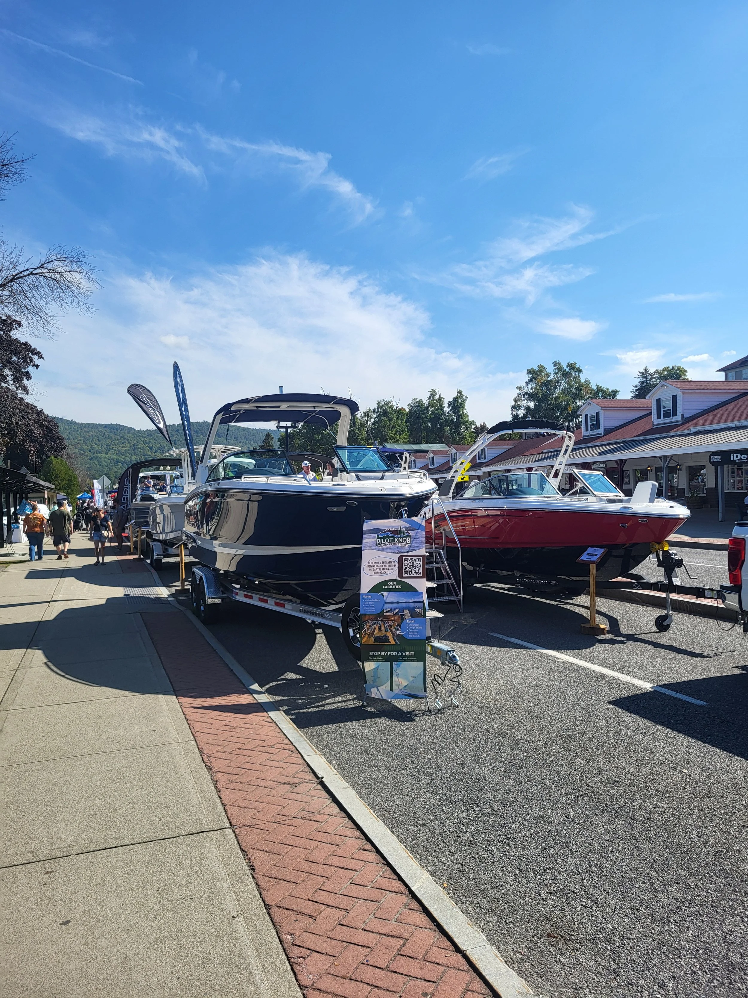 Boats on display at an outdoor boat show, parked on trailers along a sidewalk with tents and people walking around.