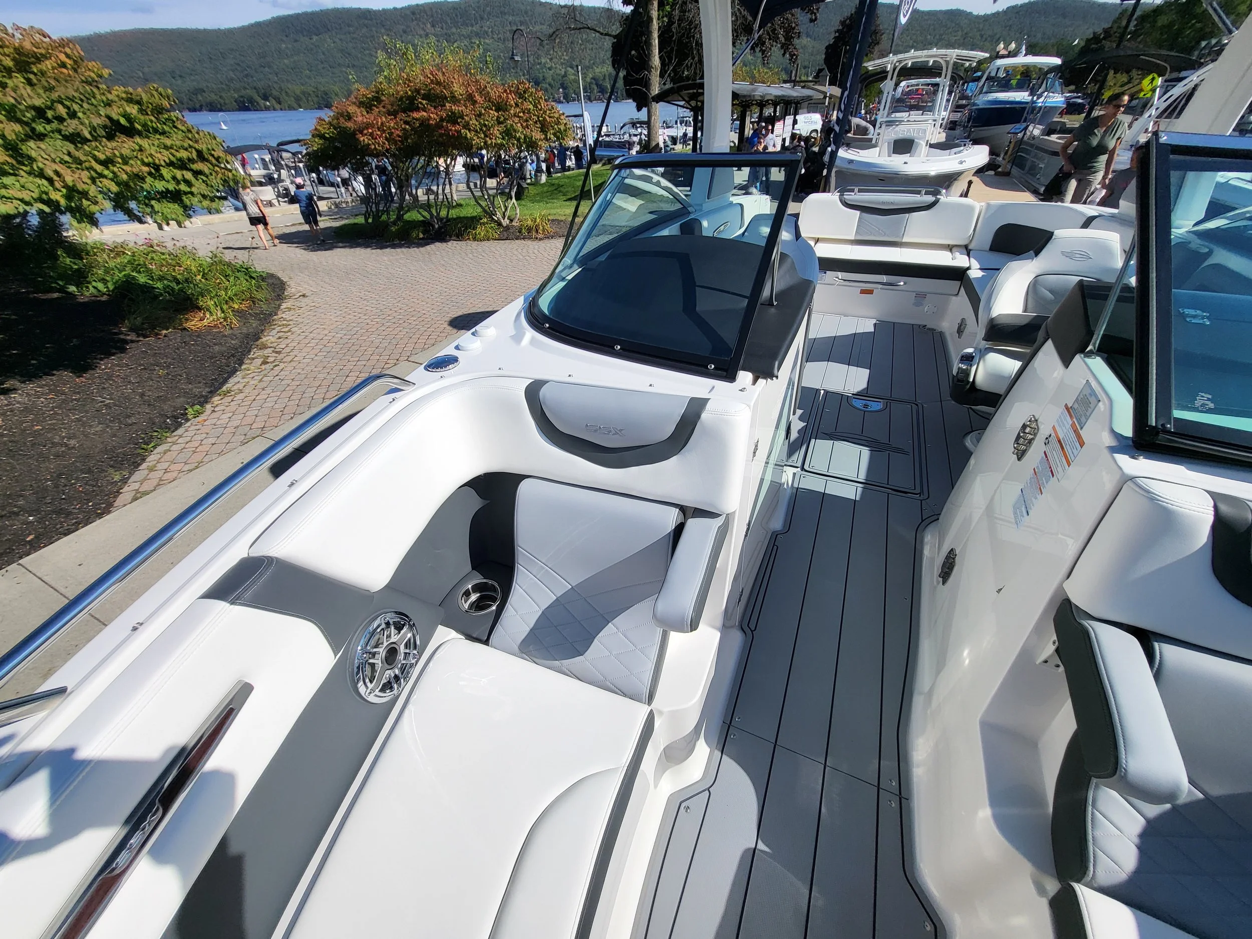 Interior of a white and gray motorboat docked at a marina, with seating and equipment visible, in a scenic area with water, trees, and people walking nearby.