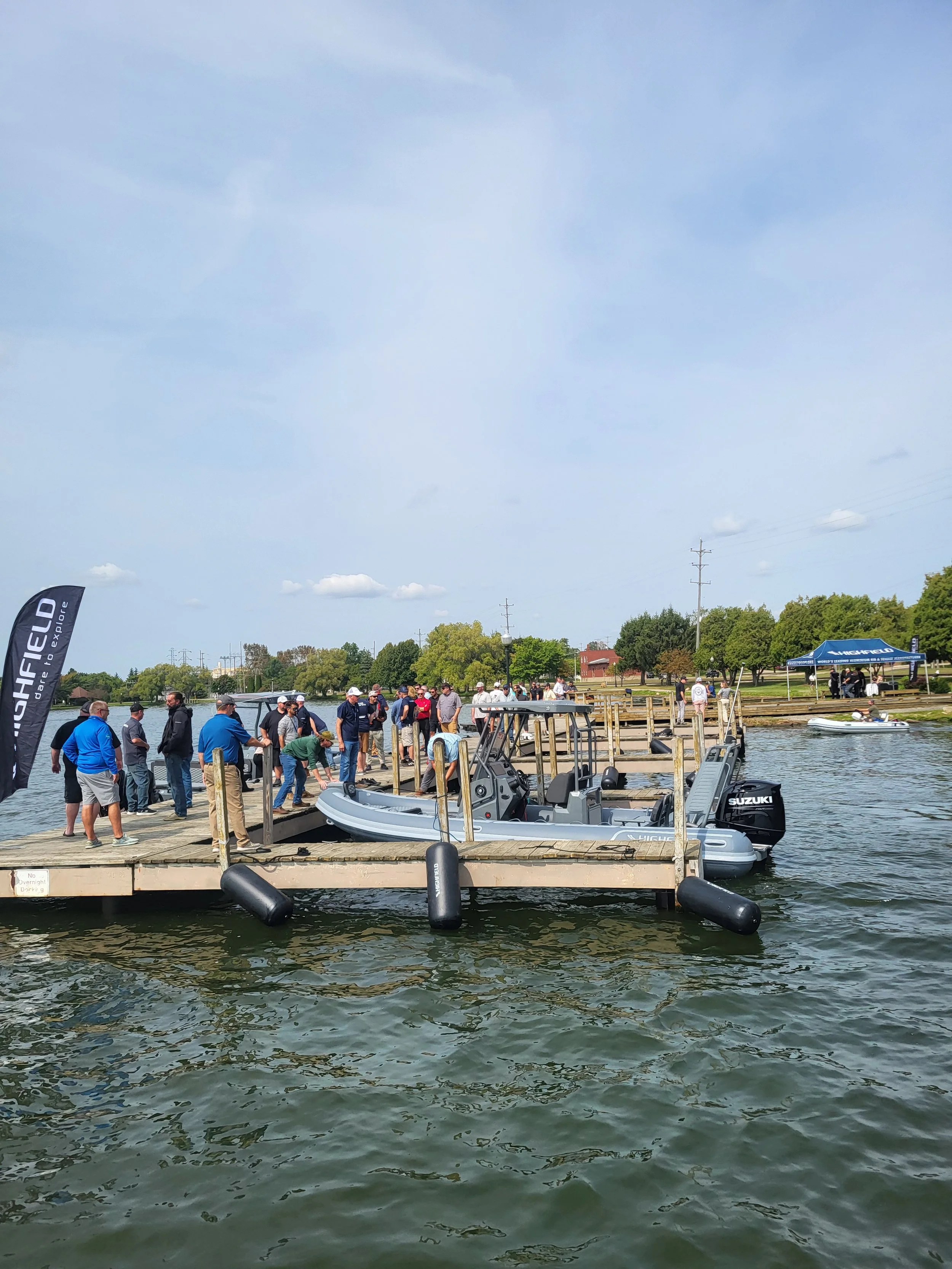 People gathered on a dock near a boat launch at a marina, preparing to launch a boat into the water, with additional boats and trees in the background.