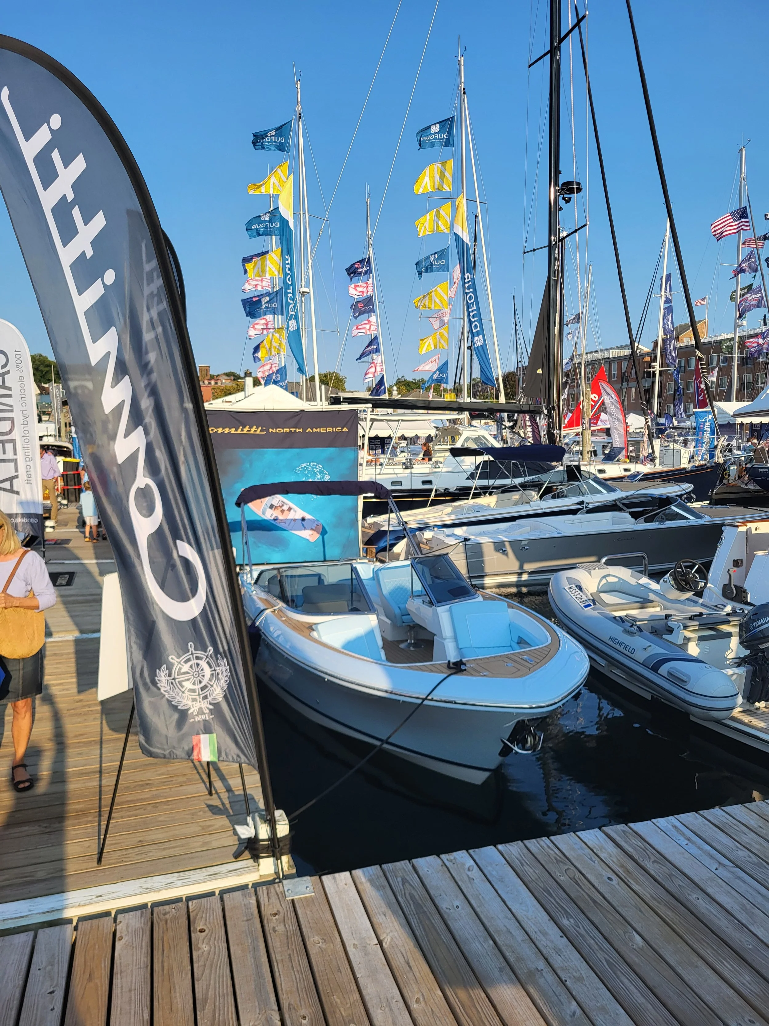 Boats docked at a marina with flags flying, people walking on the wooden pier, and banners advertising boat brands, under a clear blue sky.