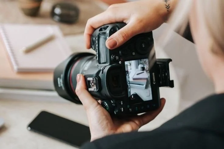 Person holding a Canon DSLR camera, taking a photo of a desk with a notebook, pen, and smartphone.