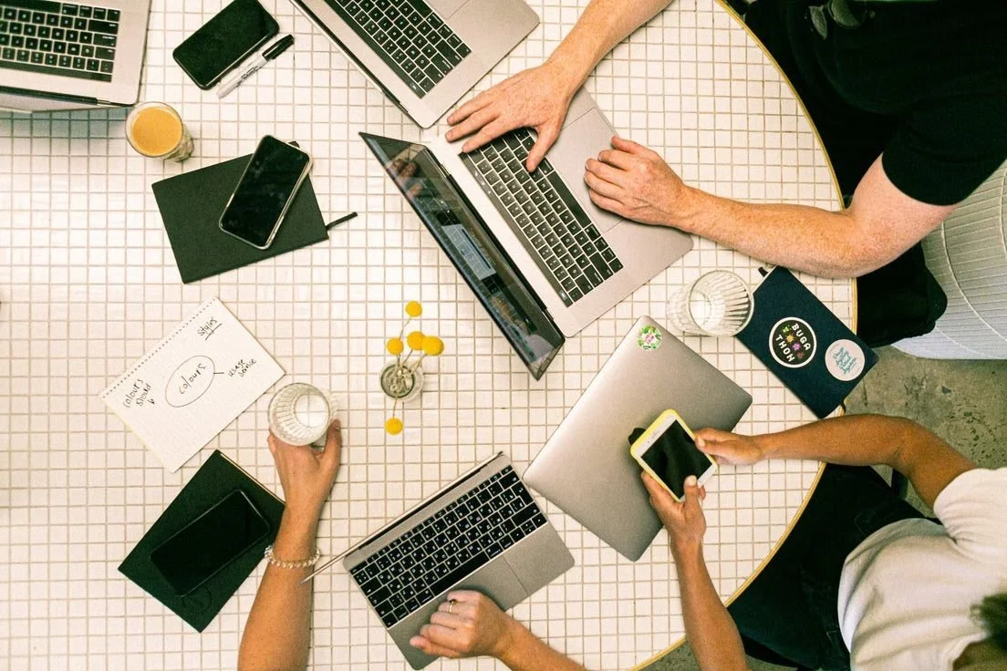 Overhead view of three people working at a round table with laptops, smartphones, notebooks, a glass of orange juice, and decorative yellow flowers.