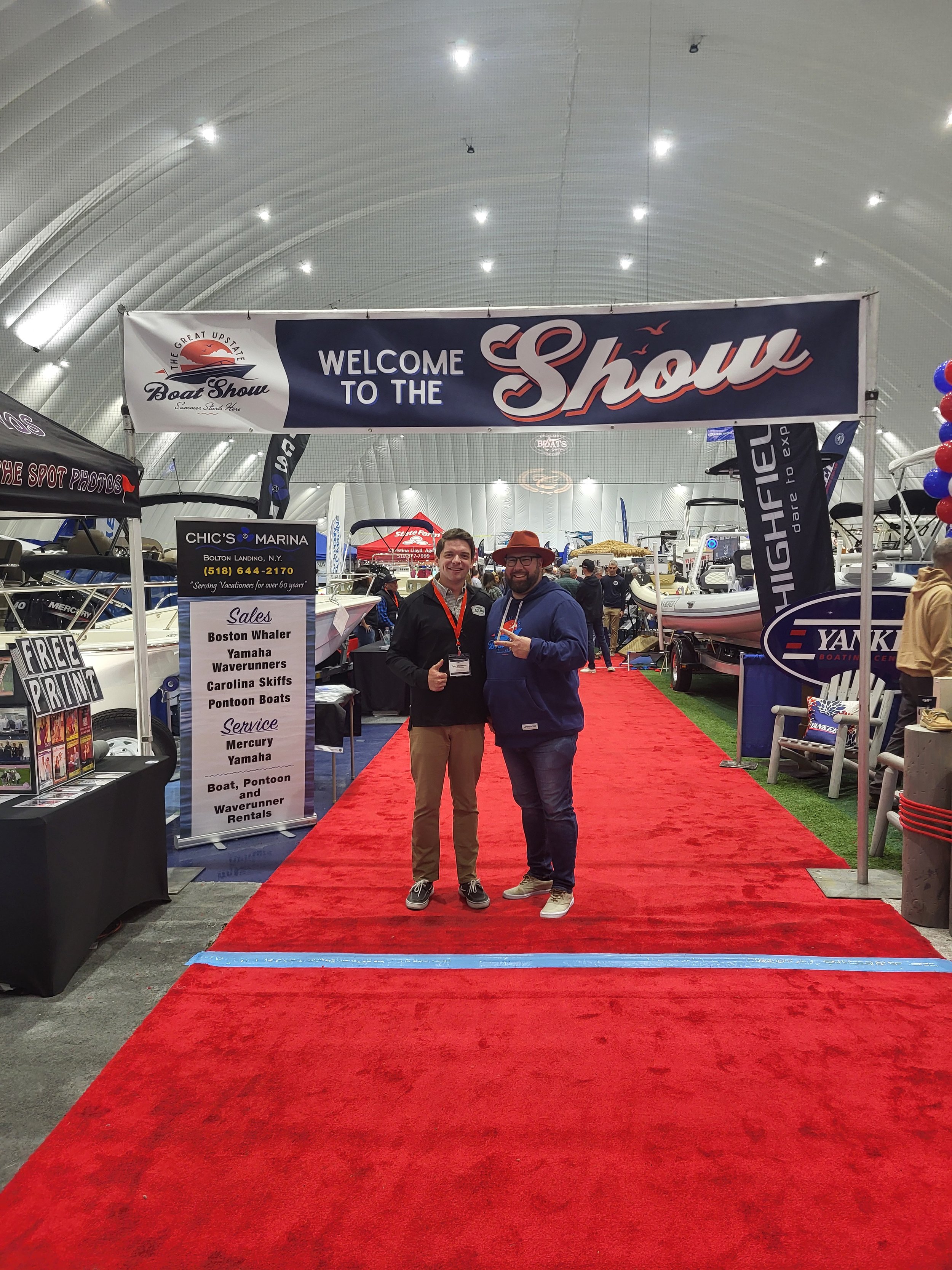 Two men standing on a red carpet inside a large boat show venue, with a large banner above them that says "Welcome to the Show." There are boats on display and various signs and booths around.