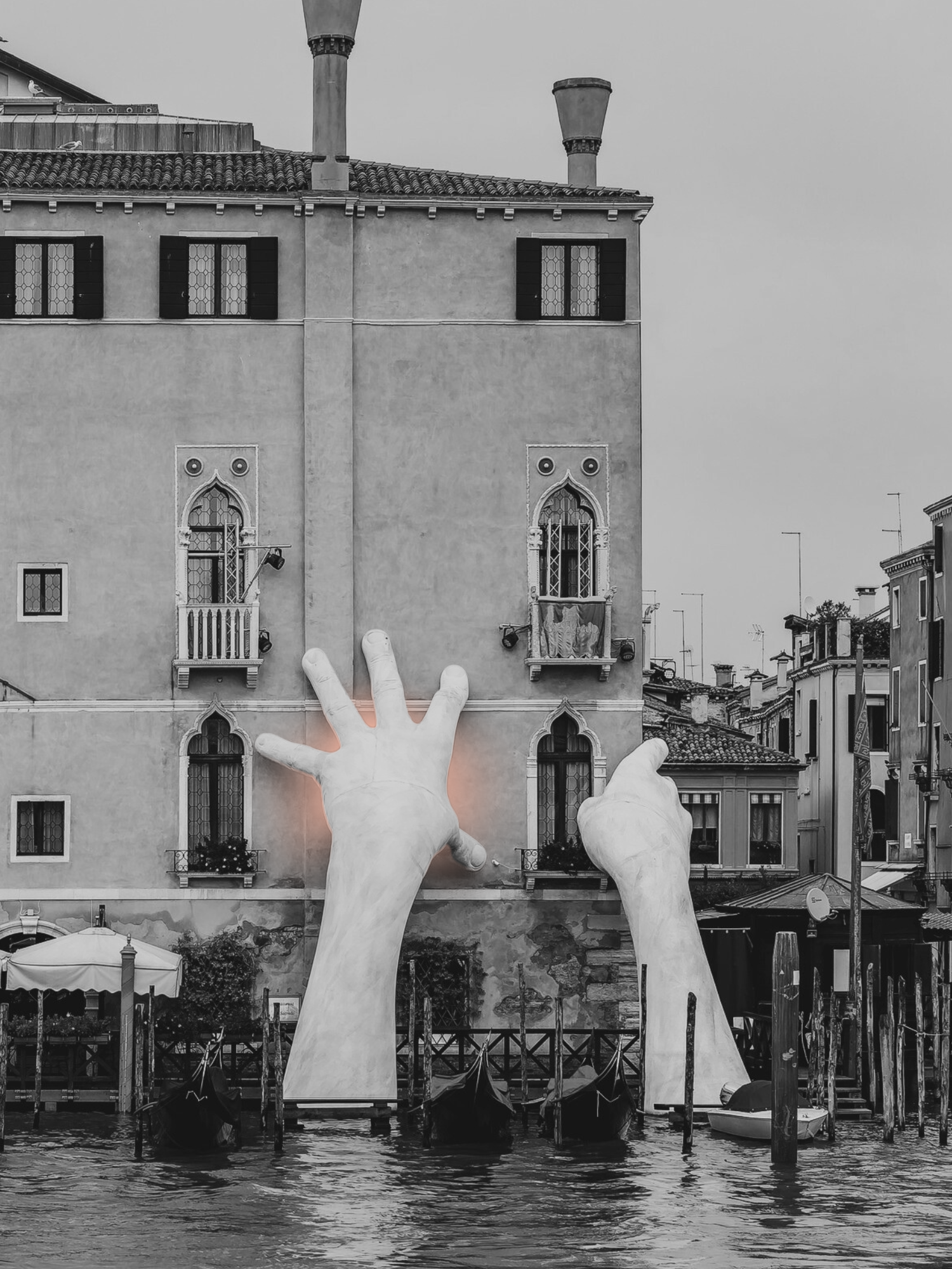 Large black-and-white photo of a building along a canal, with two large, illuminated hands reaching up from the water in front of it.