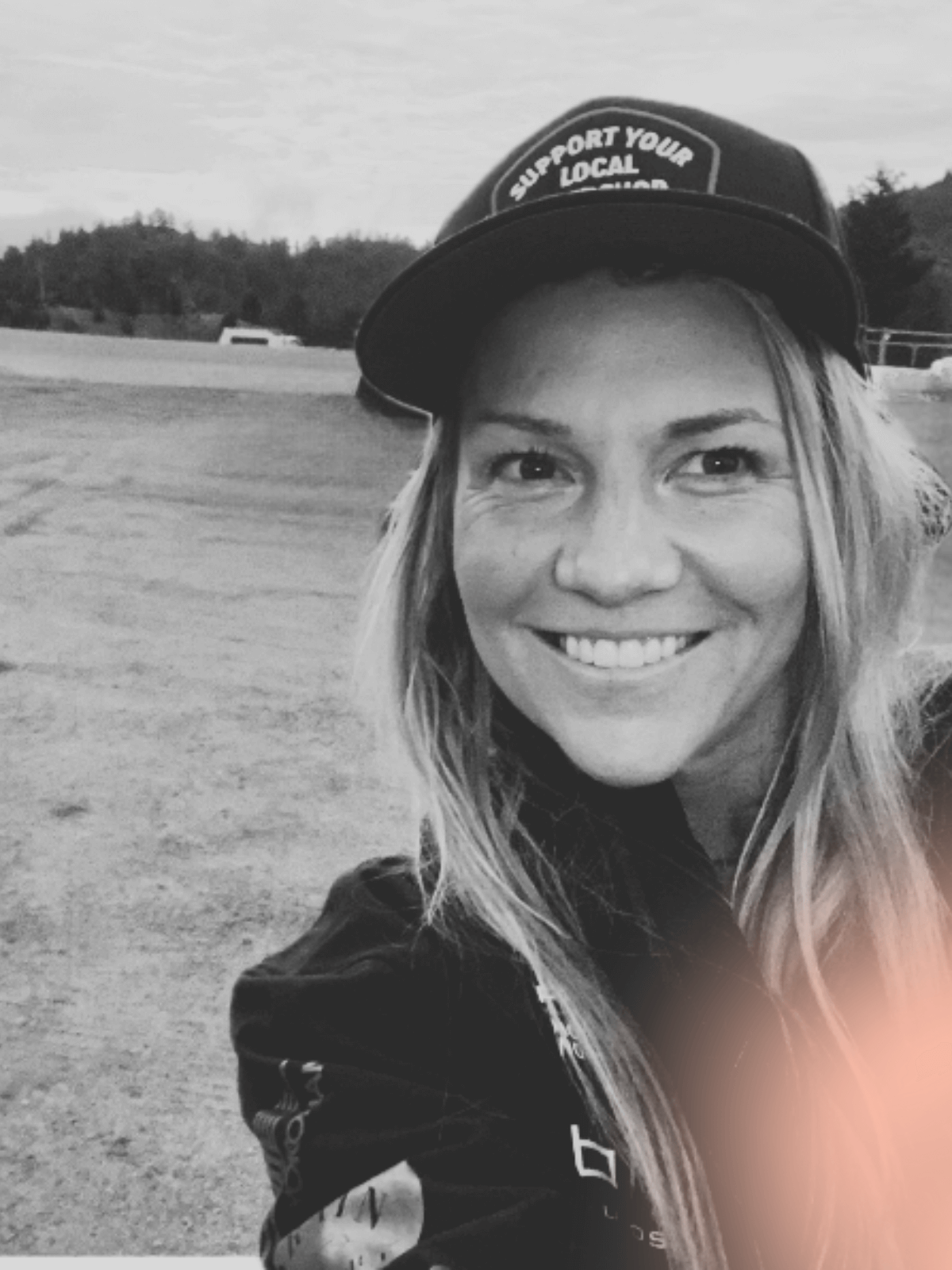 Black and white photo of a smiling woman with long hair wearing a baseball cap that reads support your local, outdoors setting in the background.
