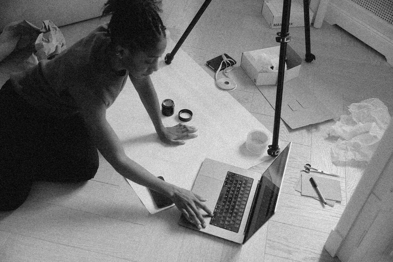 Person using laptop on the floor, surrounded by photography or artwork supplies, with a tripod and other equipment nearby.
