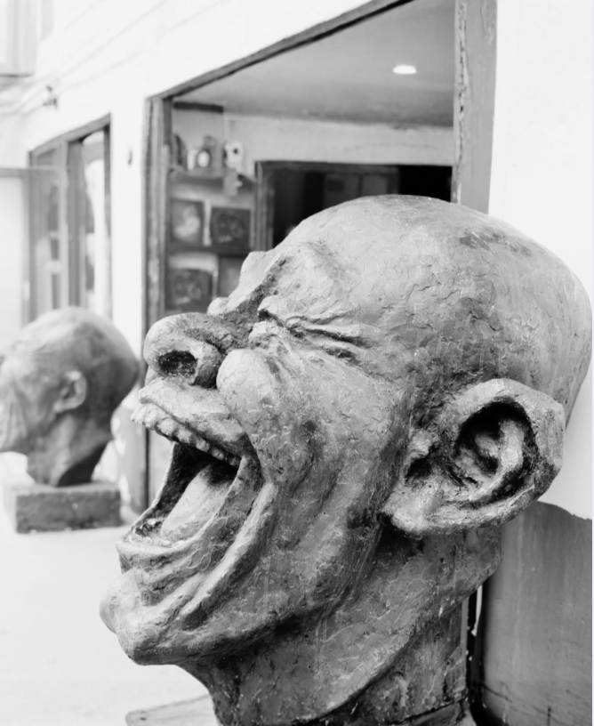 A large, distorted sculpture of a person's head with exaggerated facial features, including an open mouth, large nose, and prominent ears, positioned on a table indoors.