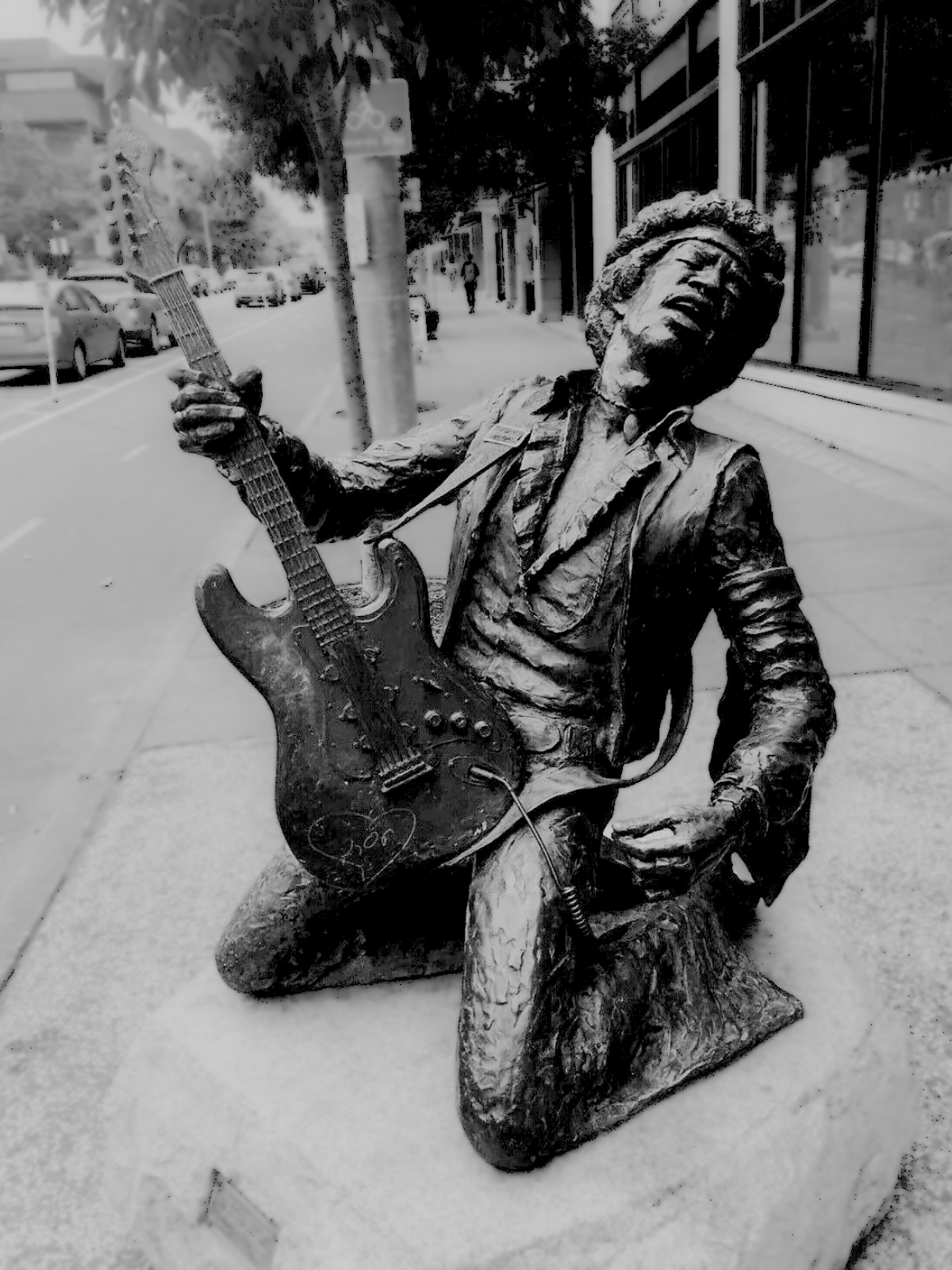 Bronze statue of a man with curly hair, kneeling on one knee, playing an electric guitar, on a city sidewalk.
