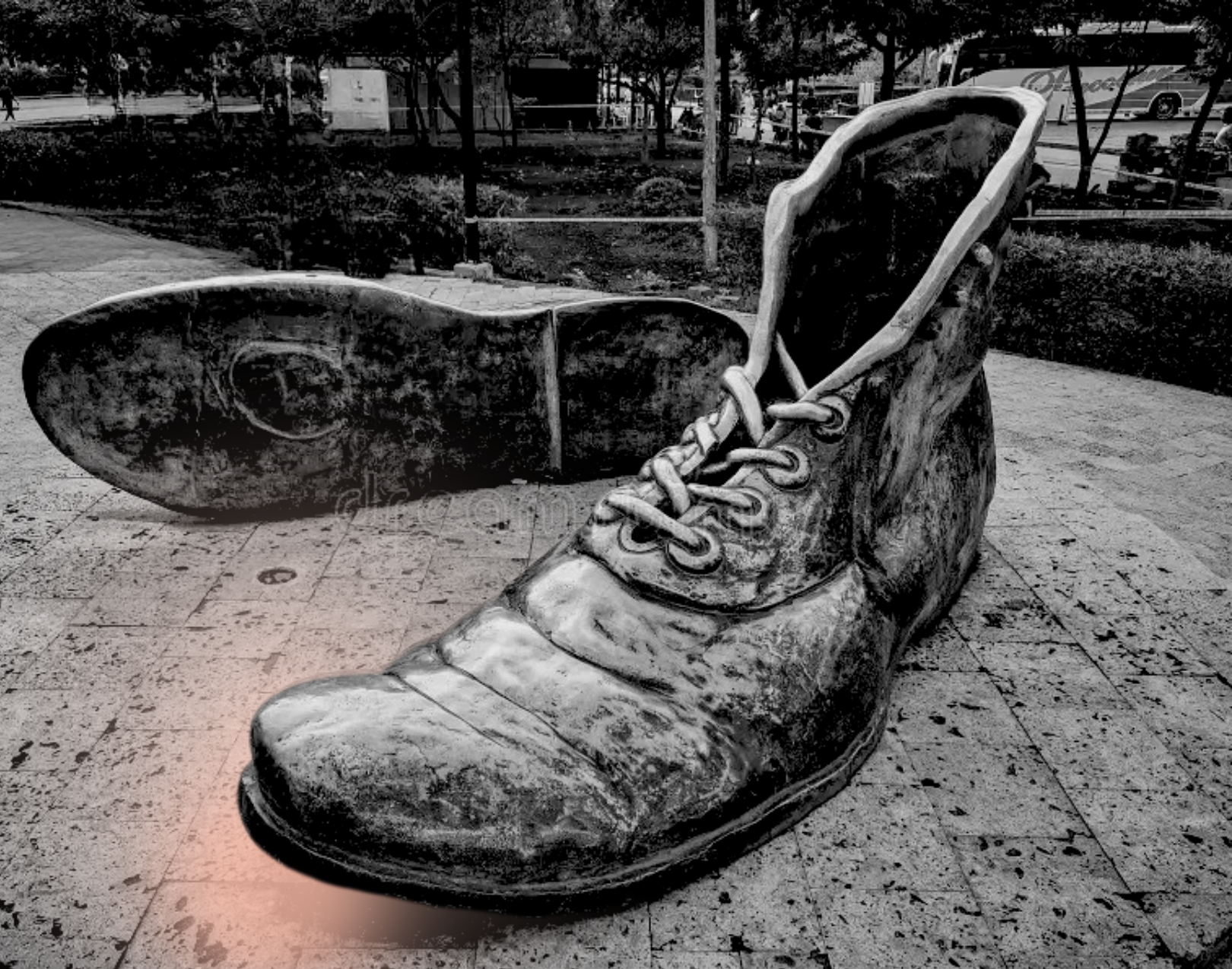 A large sculpture of a worn, old-style hiking boot with a loose shoelace, placed outdoors on a paved surface, with the sole of an oversized boot sculpture lying nearby.