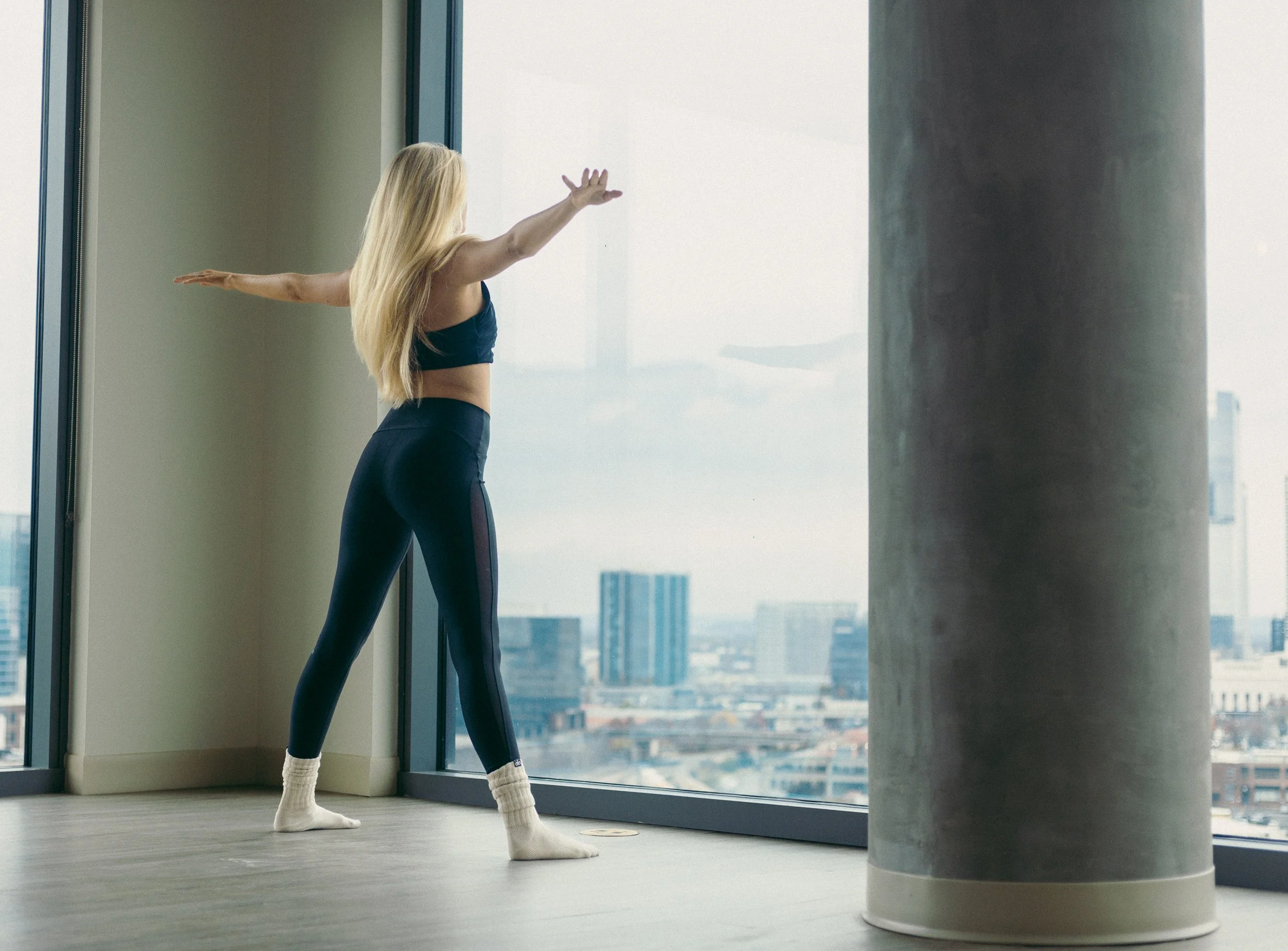 Woman in black athletic wear standing with arms extended facing Nashville skyline floor-to-ceiling window — Skyline Pilates brand shoot Alora Co Photo
