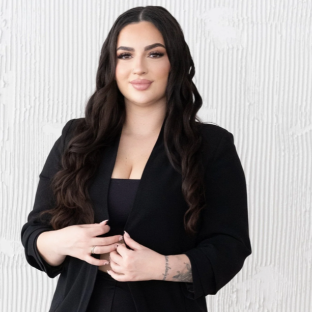 A young woman with long, wavy dark hair, wearing a black blazer and top, standing in front of a textured white wall.