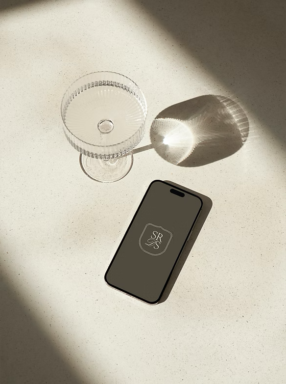 A clear, textured glass with a small amount of water, a small decorative object, and a smartphone with a logo on its screen on a light-colored surface with shadows.