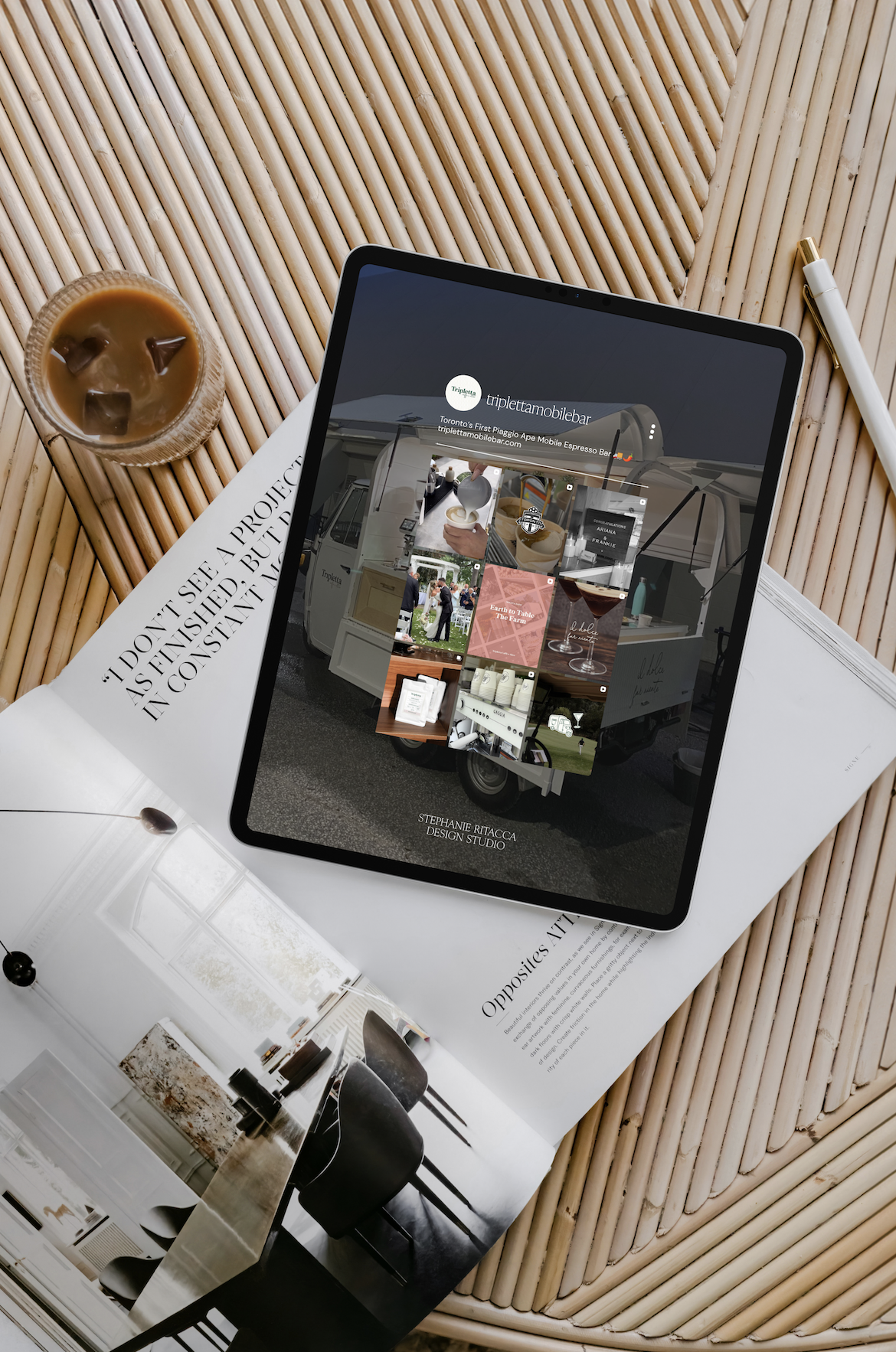 An iPad on a wooden table displaying a website for a mobile espresso bar, with a cup of iced coffee nearby, a newspaper, and an open magazine showing interior décor.