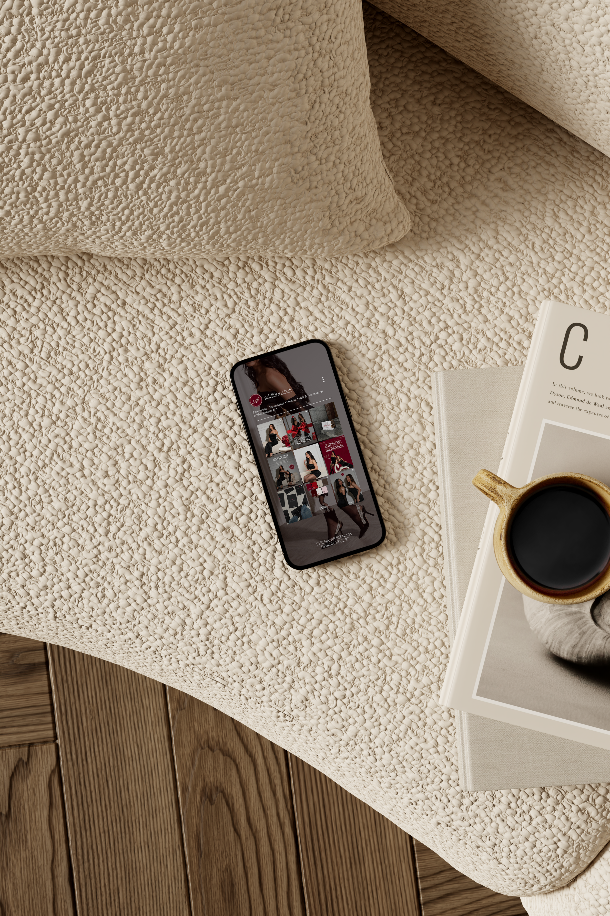 Close-up of a bed with textured bedding, a smartphone with a fashion website, a coffee mug, and books on the side table, with wooden flooring visible.