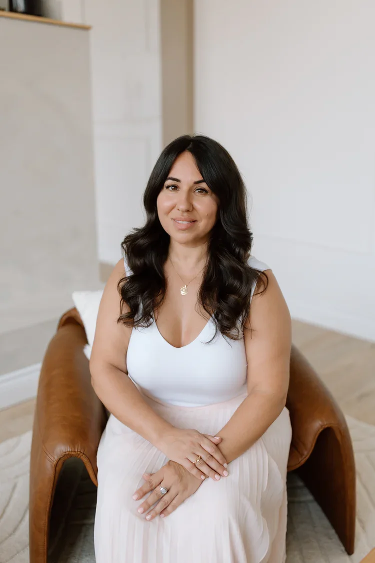 A woman with dark, wavy hair sitting on a brown leather chair in a minimalist room, smiling softly at the camera.