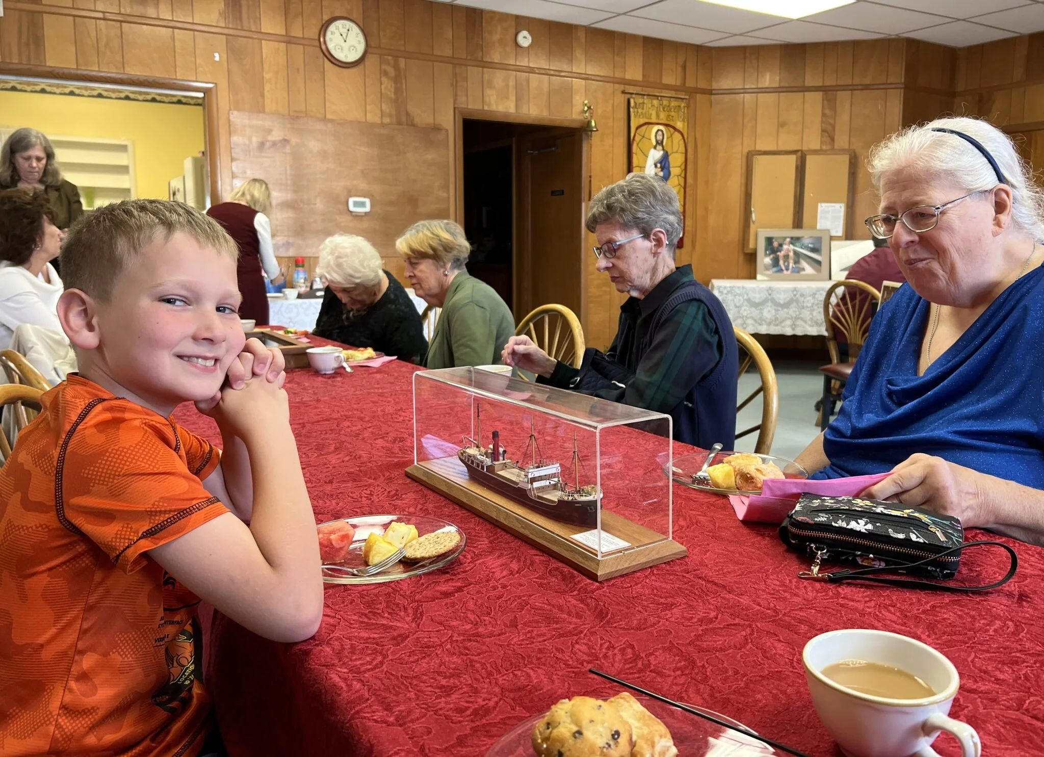 A young boy with short light brown hair sitting at a long red table with a tray of food, smiling at the camera. The table has a display of a model ship in a glass case and some food items, including a muffin and a cup of coffee. Behind him, several elderly women and men are seated, dining and engaged in conversation in a wooden-paneled room with religious artwork on the walls.