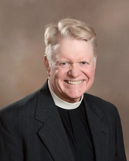 A smiling man with light-colored hair wearing a clerical collar and a dark suit against a neutral background.