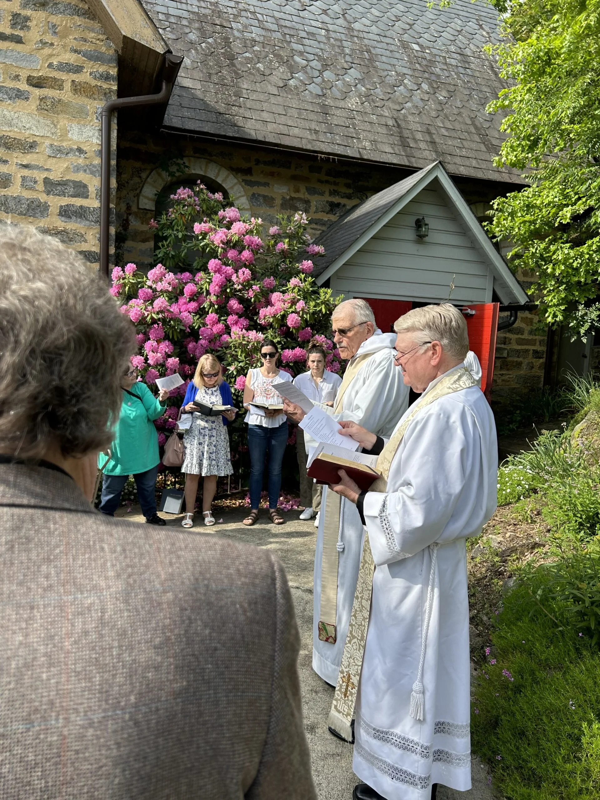 People participating in an outdoor religious ceremony or prayer, with two priests and several congregants standing near a stone building with pink flowering bushes and greenery.