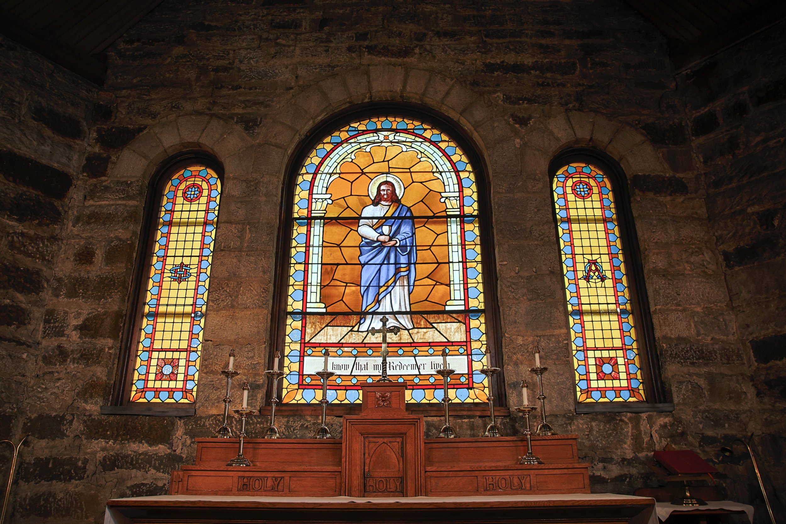 Stained glass window in a church depicting Jesus Christ with a halo and holding a cup, with two smaller colorful windows on either side, behind an altar with candles and religious symbols.