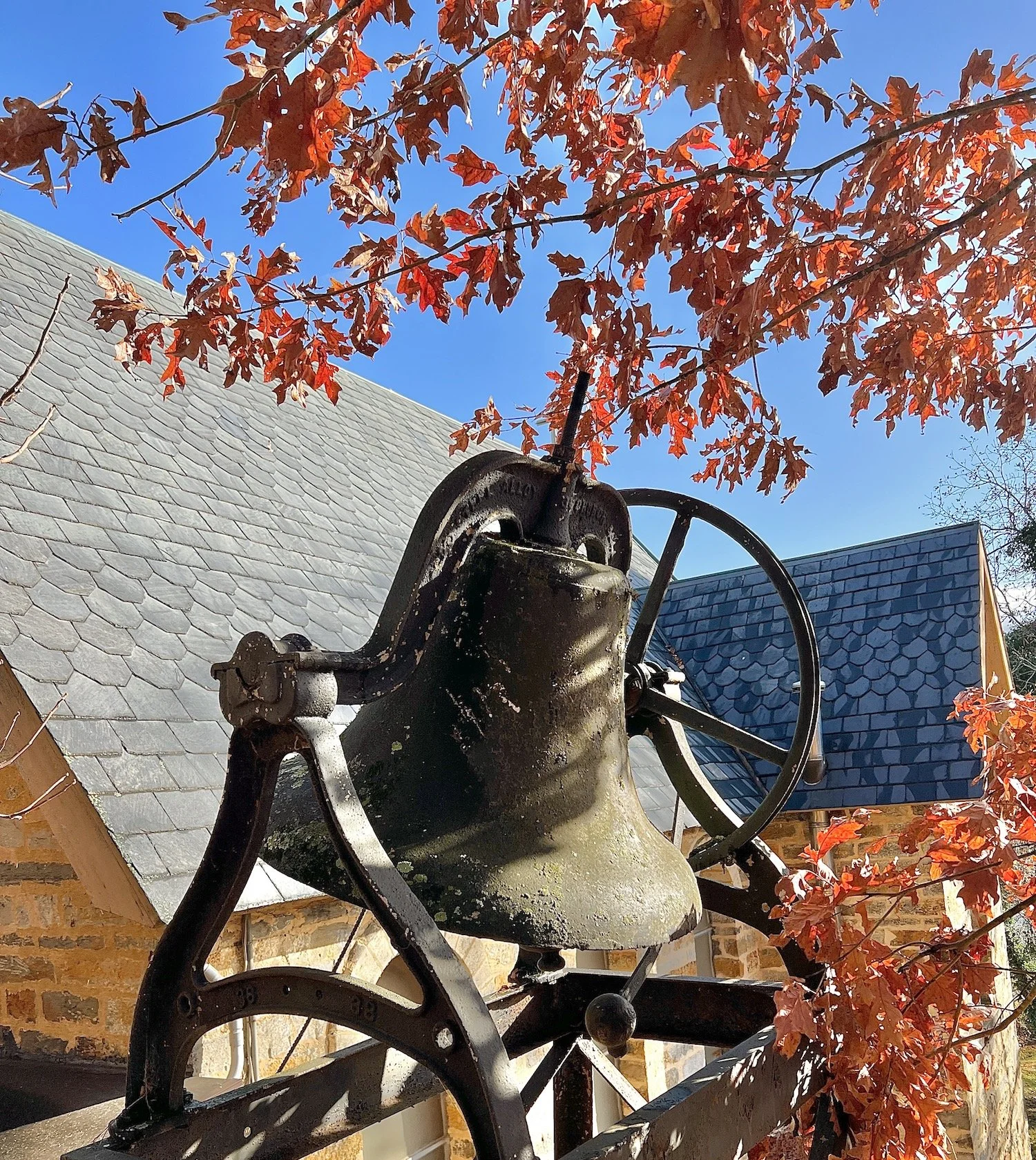 An old outdoor bell mounted on a metal frame, with red autumn leaves and a blue sky in the background.