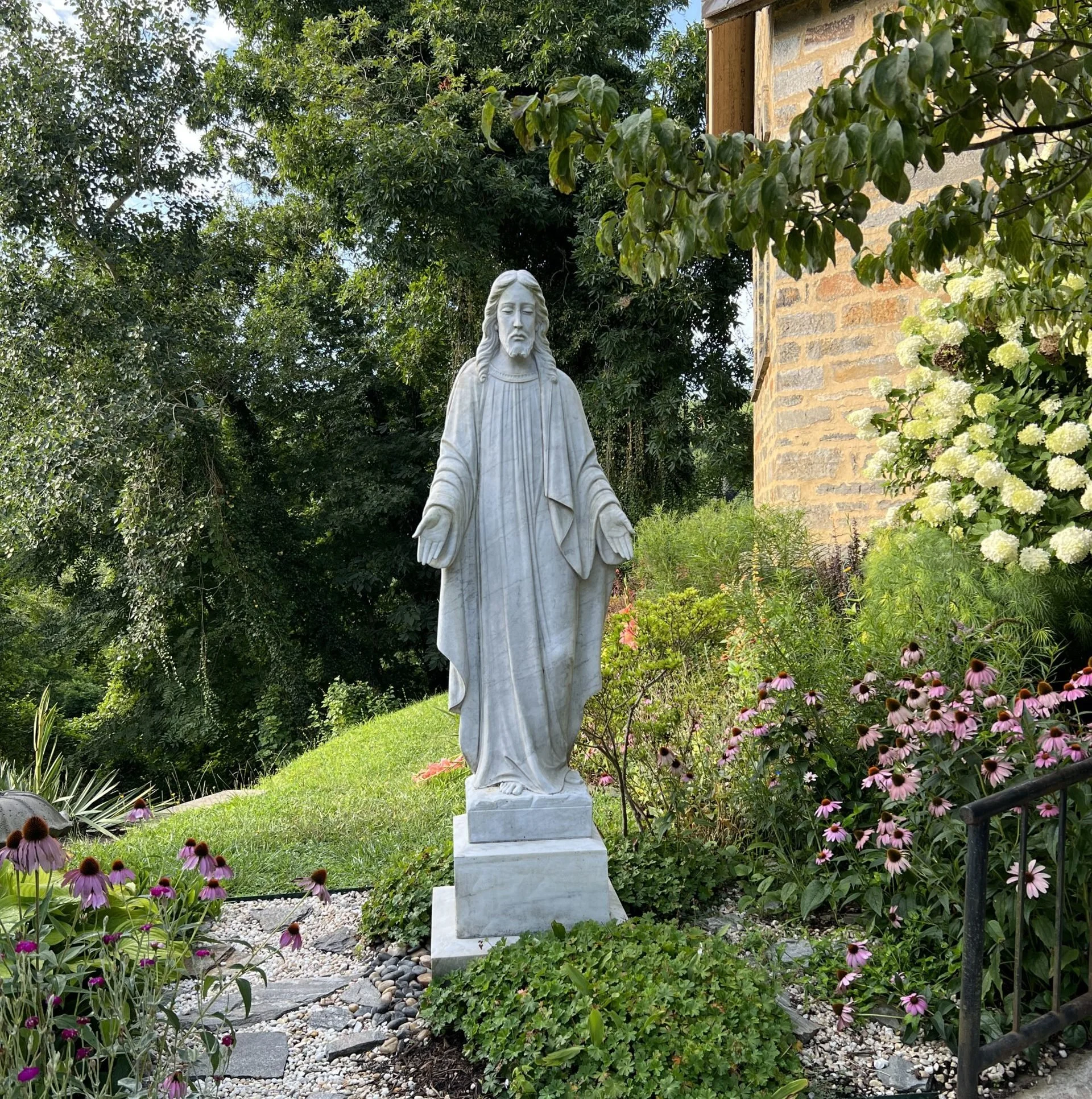 A white marble statue of Jesus Christ standing in a lush garden with green trees, pink and white flowers, and a brick building in the background.